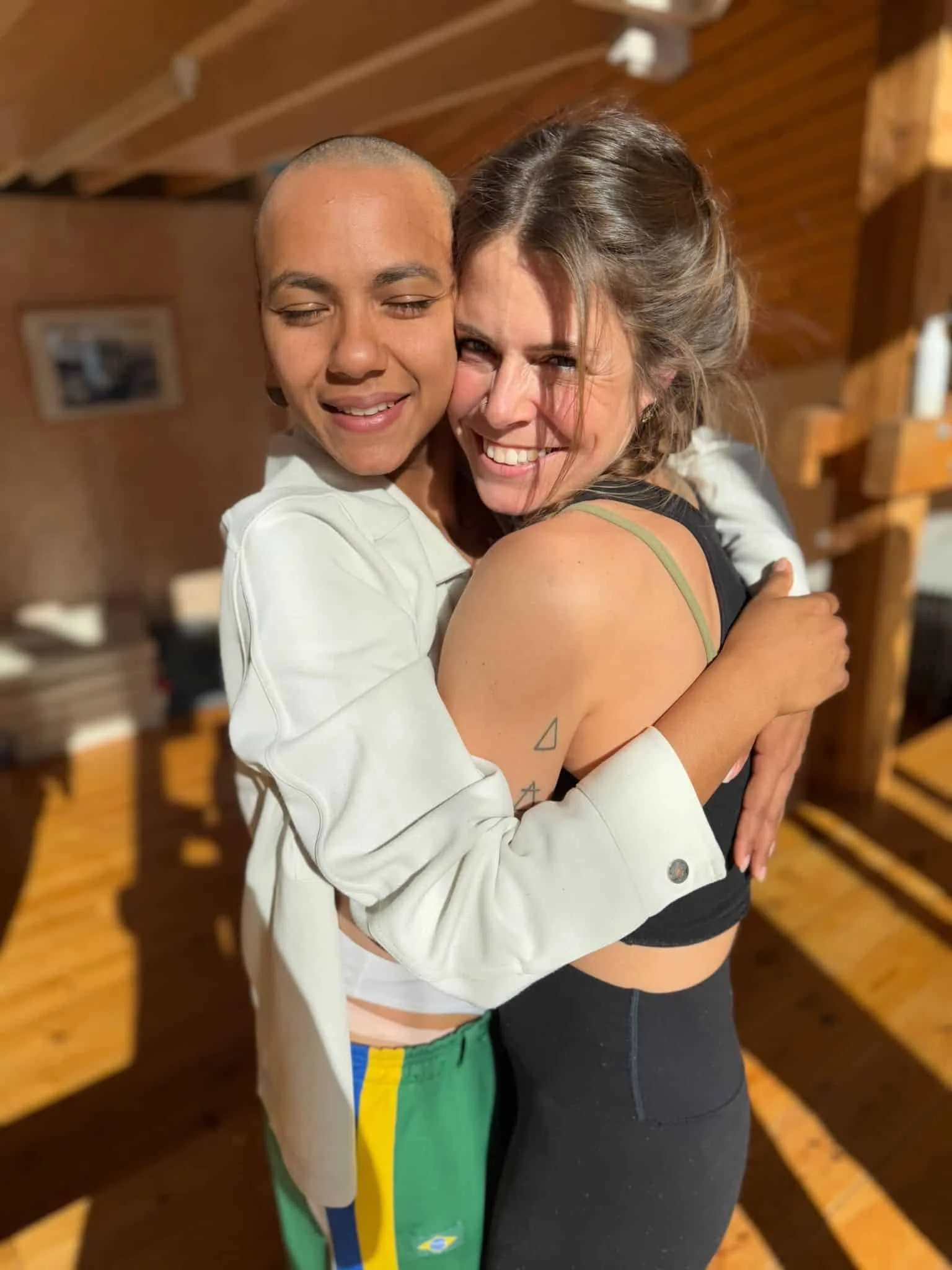 Two women hugging, one with a shaved head and brown skin, the other with brown hair and slightly darker skin, smiling with eyes closed, in a cozy indoor setting with wooden walls.