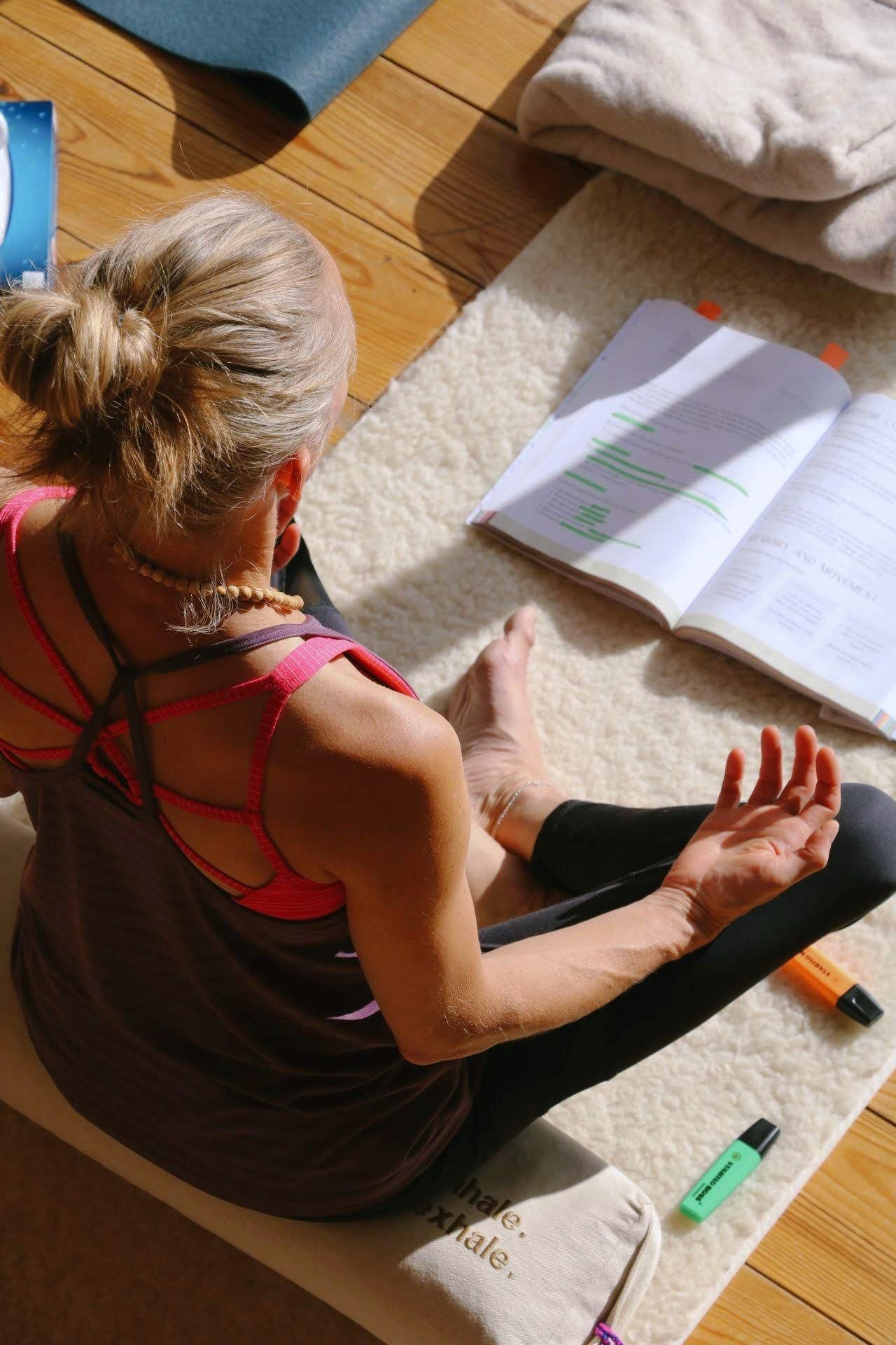 A woman sitting cross-legged on the floor, reading or studying with an open book in front of her. She has her hair in a bun, is wearing a sleeveless top, and surrounded by highlighters and a notebook.