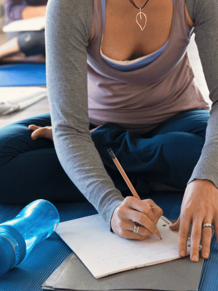 A woman practicing yoga on a blue mat, writing notes in a notebook with a pencil, with a water bottle nearby.
