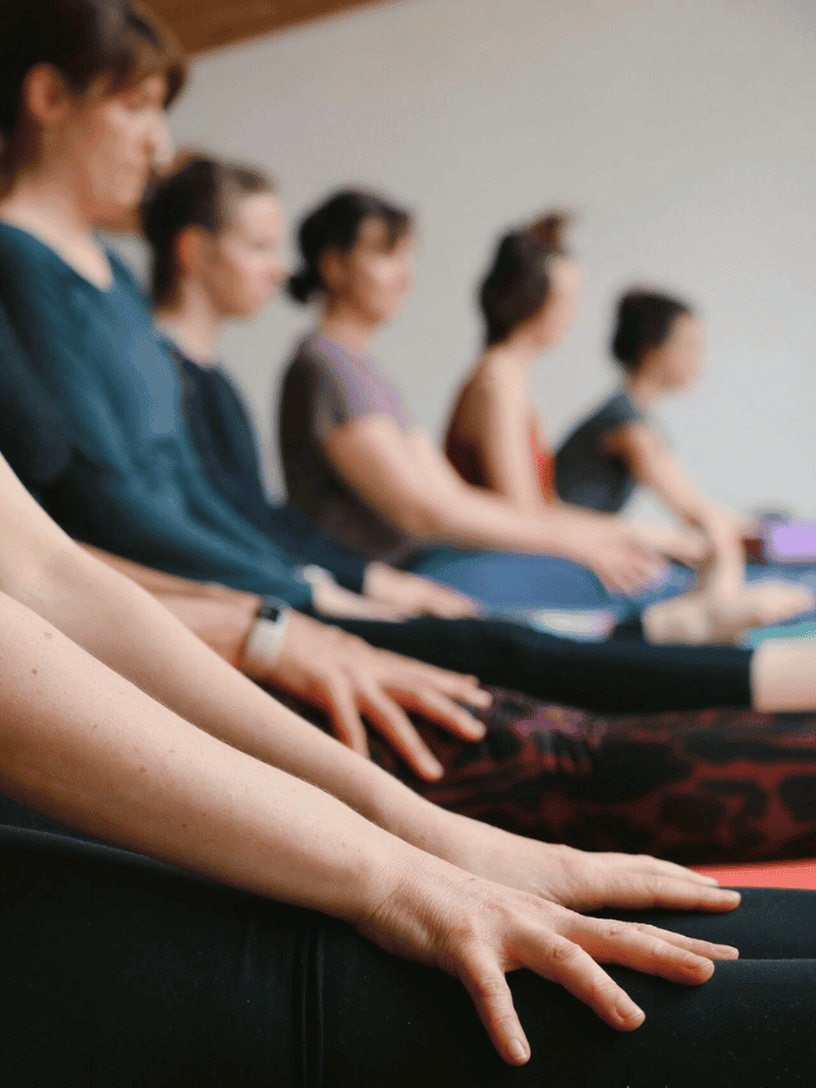 Close-up of people sitting cross-legged in a yoga or meditation class, focusing on their hands resting on their knees.