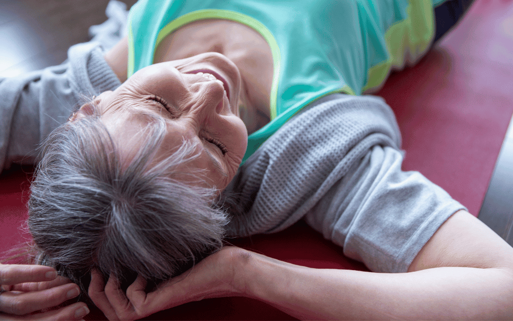 An elderly woman lying on a bed, smiling with eyes closed, and resting her head on her arm.