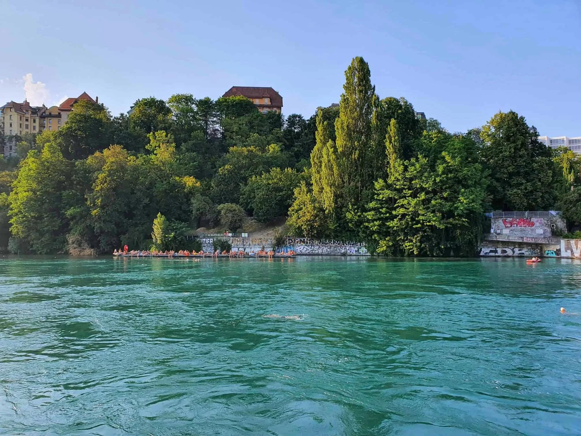 A river in Geneva with turquoise water, a line of people on the shore, some swimming and a small boat in the water, with green trees and buildings on a hill in the background under a clear blue sky.
