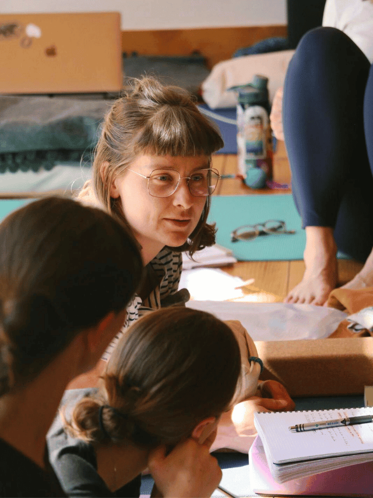 A woman with glasses and striped shirt talking to two people with brown hair, in a room with notebooks, a pen, and various personal items on a table.