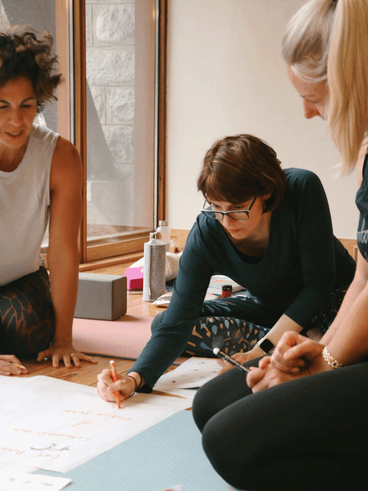 Three women are working together on a large sheet of paper on a wooden floor, brainstorming or planning, with various supplies nearby.
