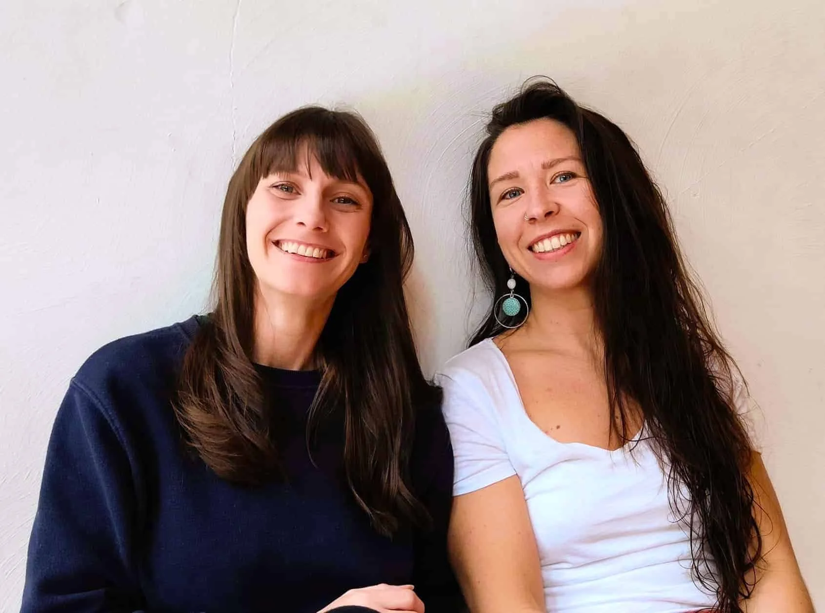 Two women smiling and sitting against a plain white wall, one with brown hair in a black shirt, and the other with long dark hair and turquoise earrings, wearing a white top.