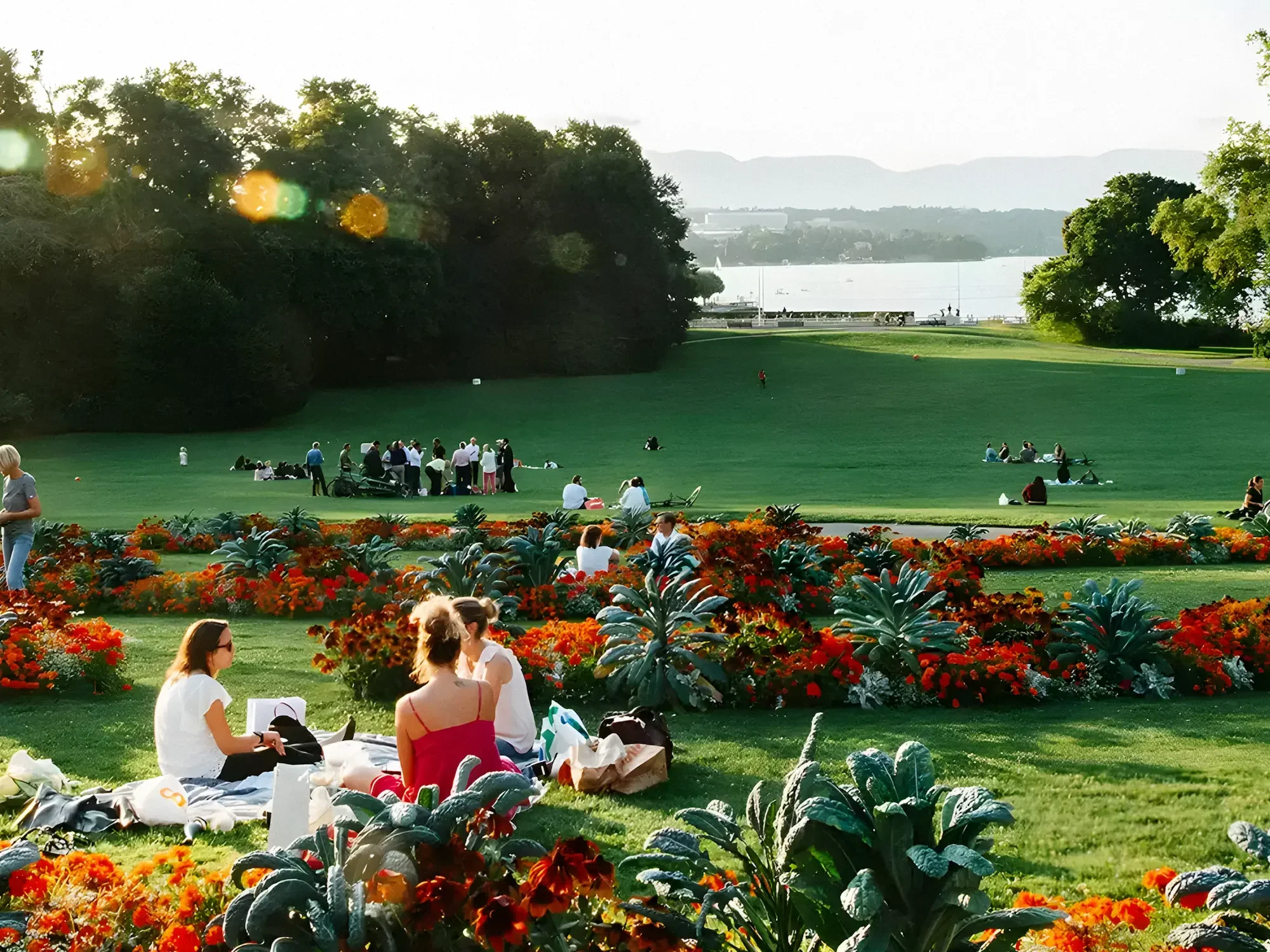 People having picnics and relaxing in a park in Geneva with colorful flowers, lush green grass, trees, and a body of water in the background.