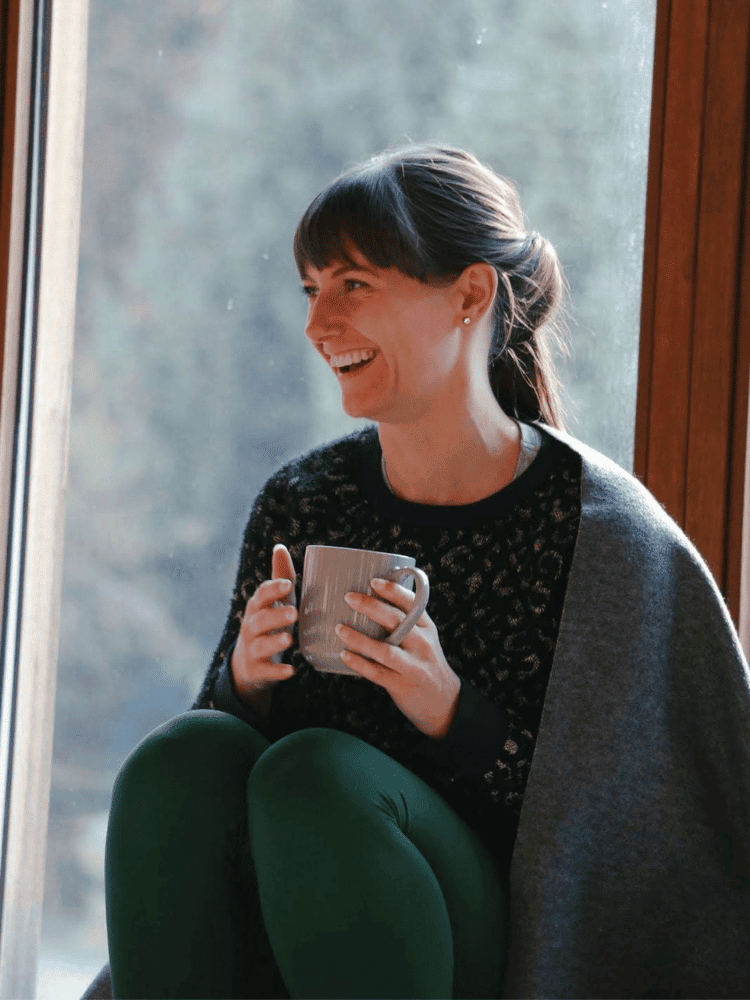 A woman sitting by a window, smiling and holding a mug, wearing a black sweater and green yoga pants.