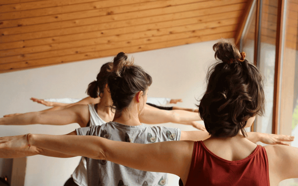 Three women in a yoga class stretching with arms extended outward inside a room with a wooden ceiling and a large window.