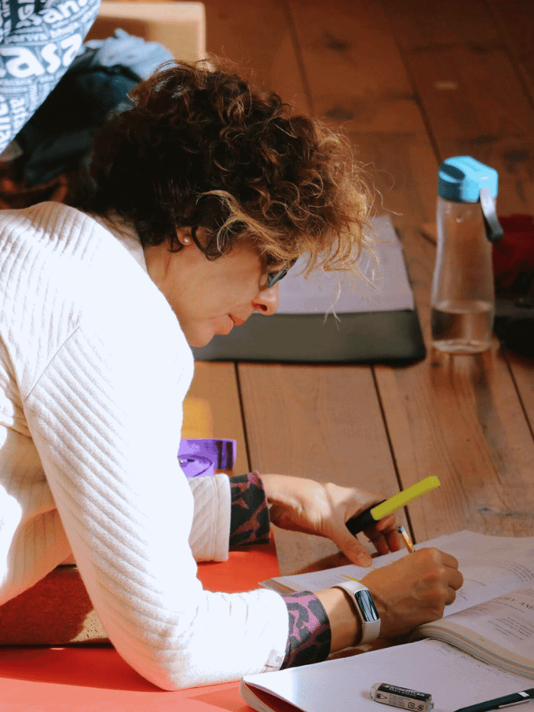 Woman with curly hair and glasses reading and writing in a notebook on a wooden floor, with a water bottle and backpack nearby.