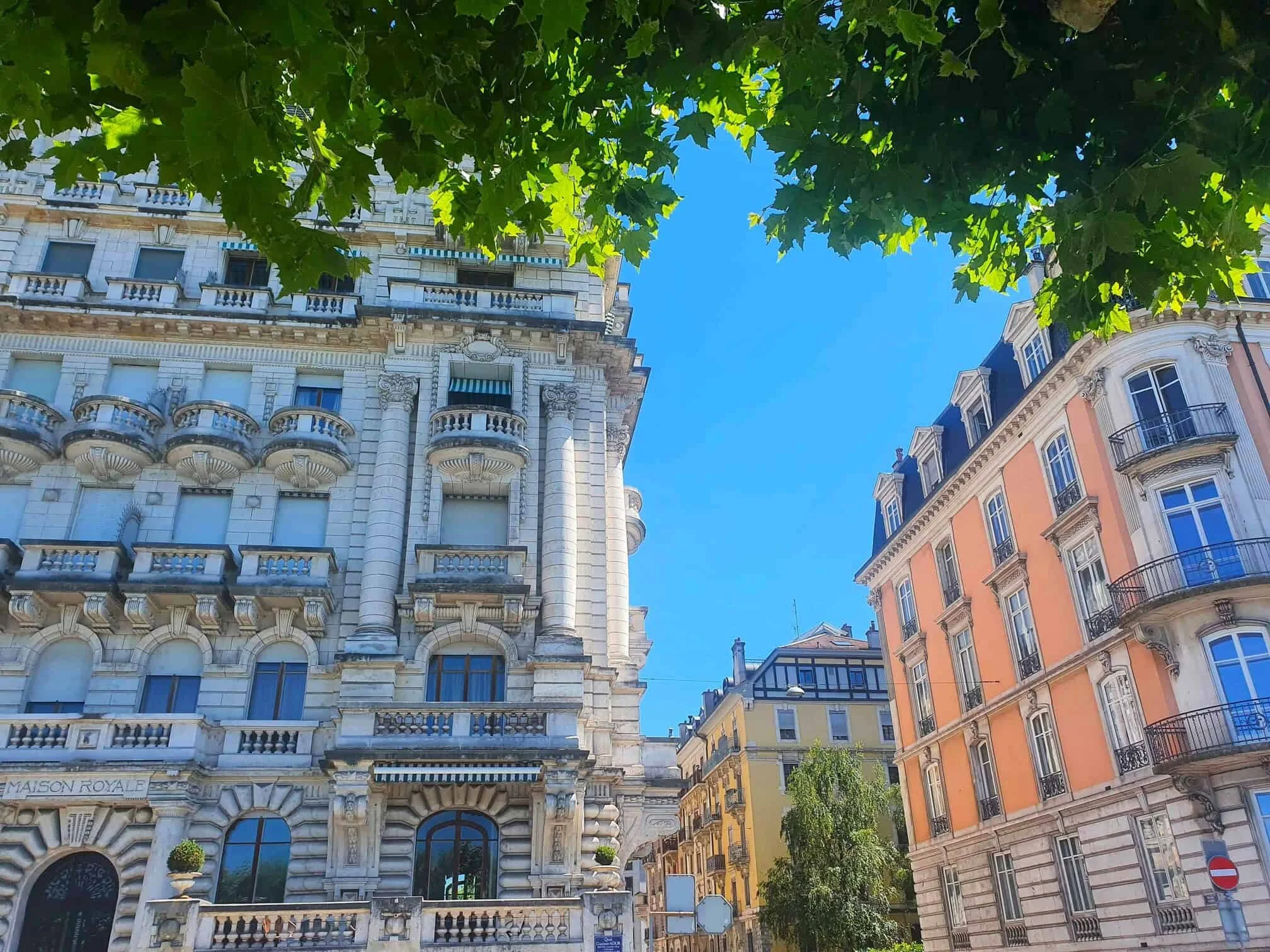 Street view of historic European-style buildings with ornate facades, balconies, and a clear blue sky. Tree branches frame the top of the image. Eaux Vives Geneva.