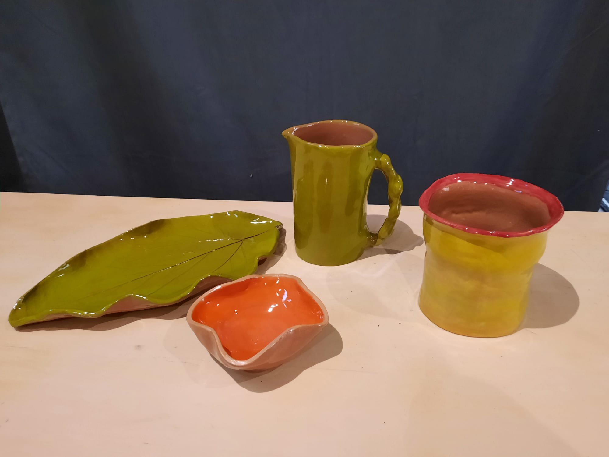 Set of handcrafted ceramic pottery including a yellow mug with a handle, a matching yellow bowl with a red rim, a yellow leaf-shaped plate, and a small orange bowl, all placed on a light-colored surface against a dark background.