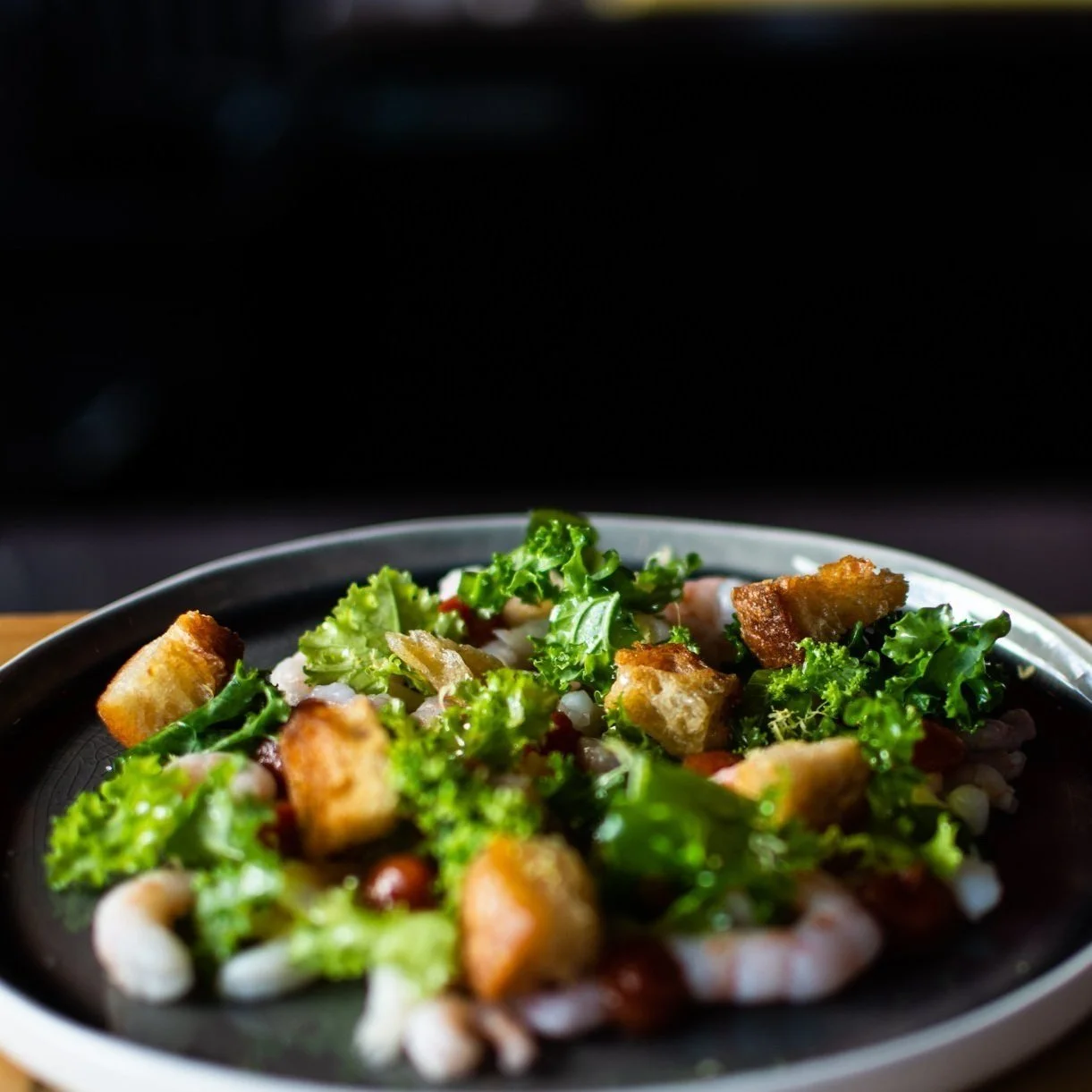 A plate of seafood salad with shrimp, sourdough croutons, kale, black truffle and cocktail sauce on a black plate.