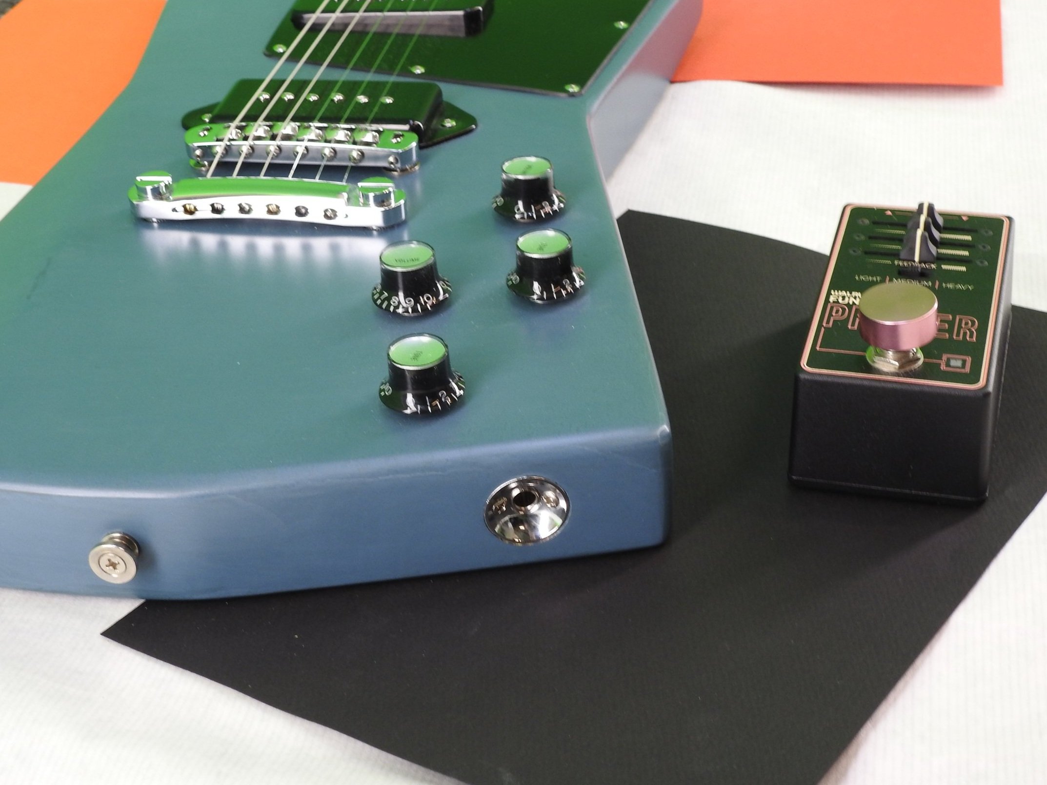 Close-up of a vintage-style guitar with a blue body, black knobs, silver hardware, and a green pickguard, placed on a multicolored surface.