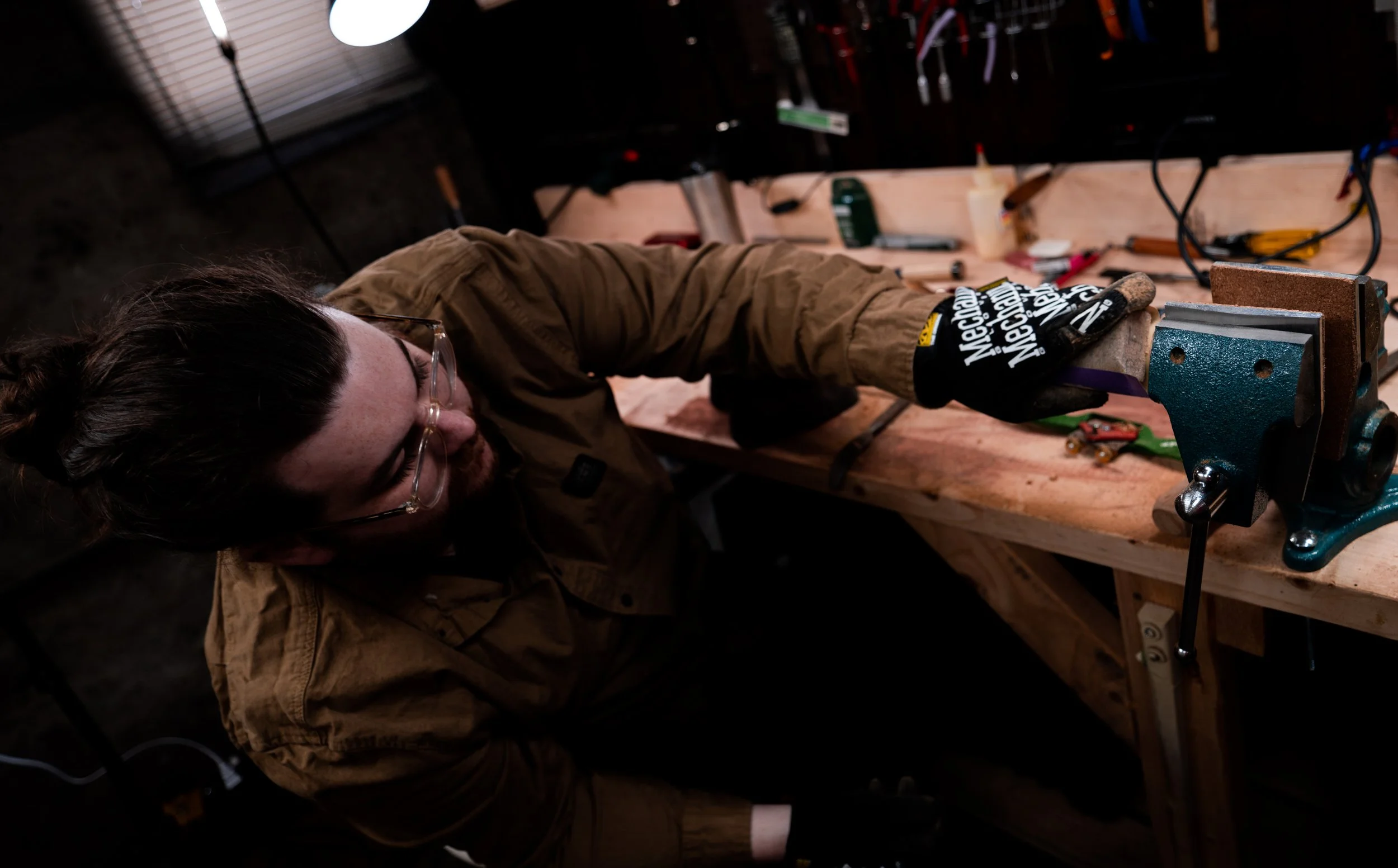 A man working at a woodworking bench, wearing safety glasses and gloves, with tools and equipment on the work surface.