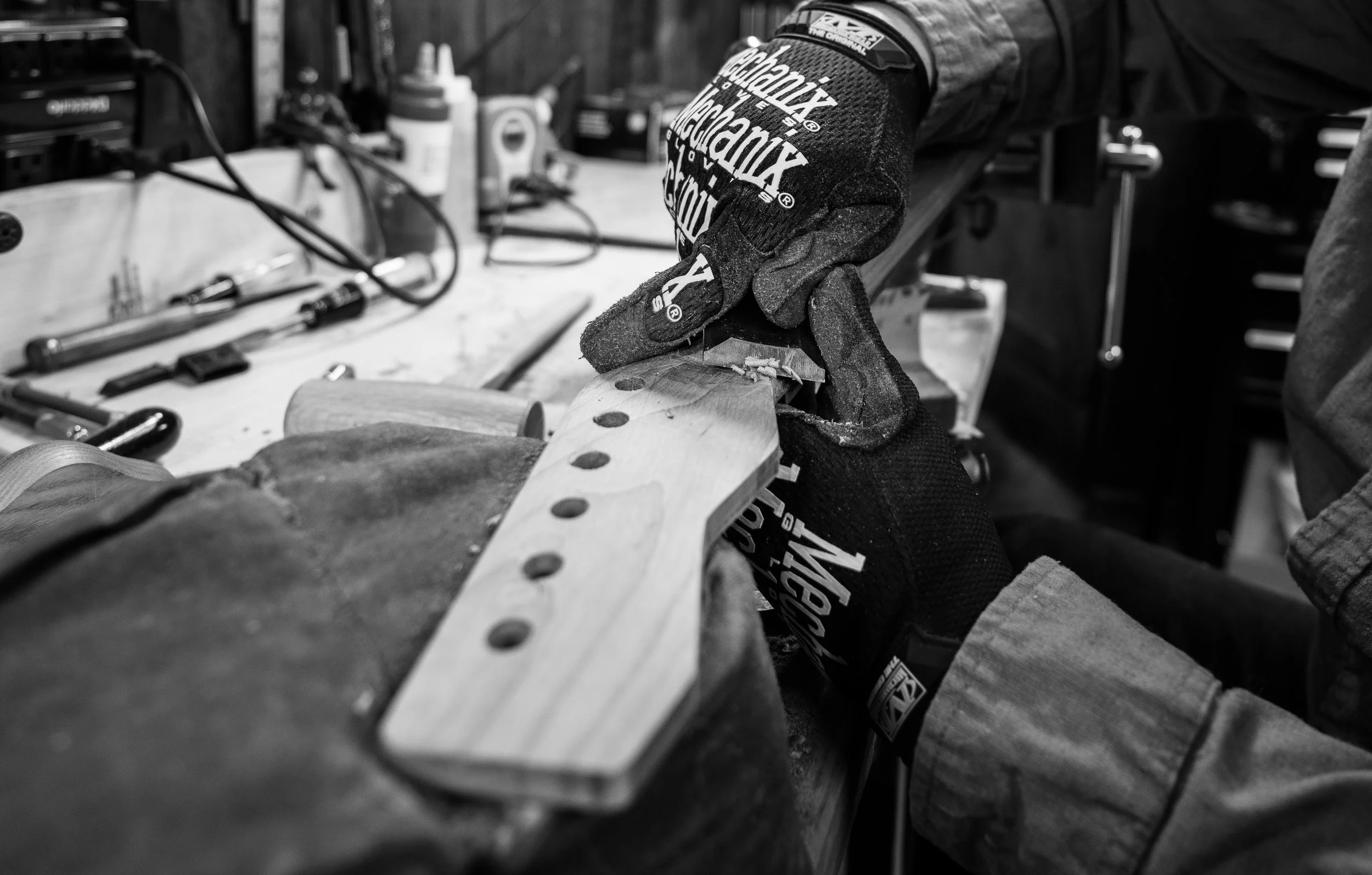 A person wearing gloves working on a guitar neck, sanding or shaping the wood, in a workshop with various tools and supplies in the background.