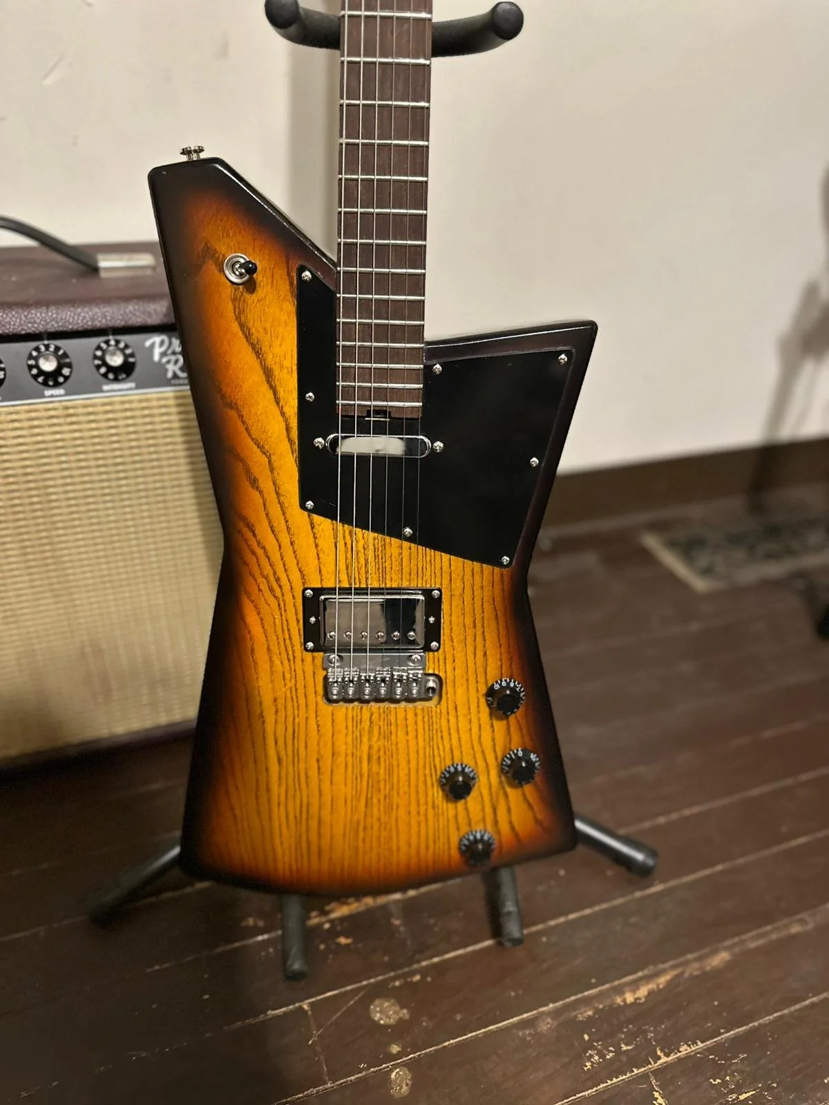 A unique electric guitar with a triangular body shape and a sunburst finish, standing on a guitar stand in front of a small amplifier.