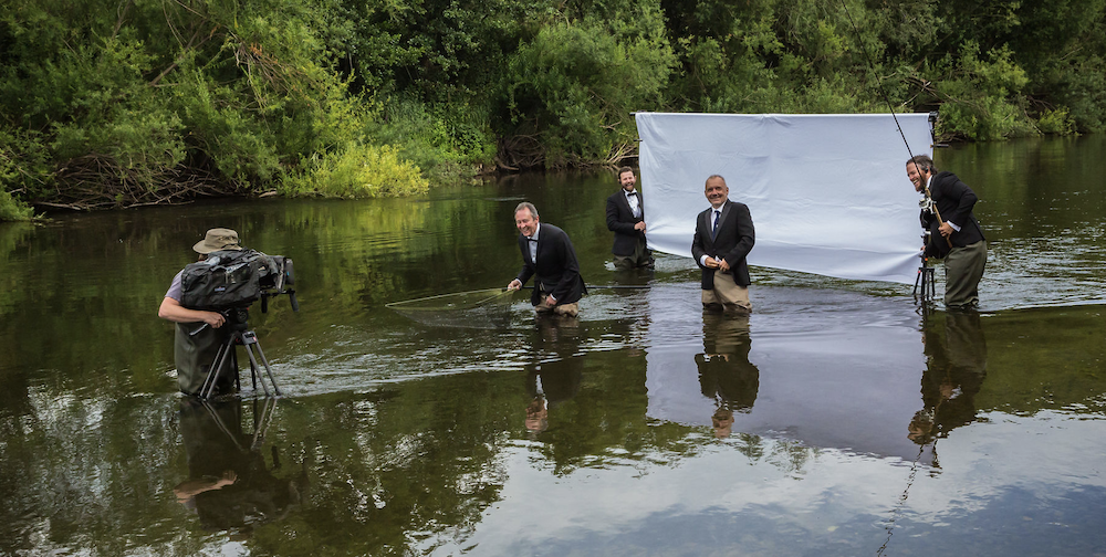 A camera crew films four men in suits standing in a river laughing