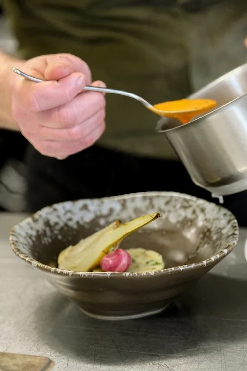 Close up of a stone bowl being plated up by a chef with a bright orange sauce