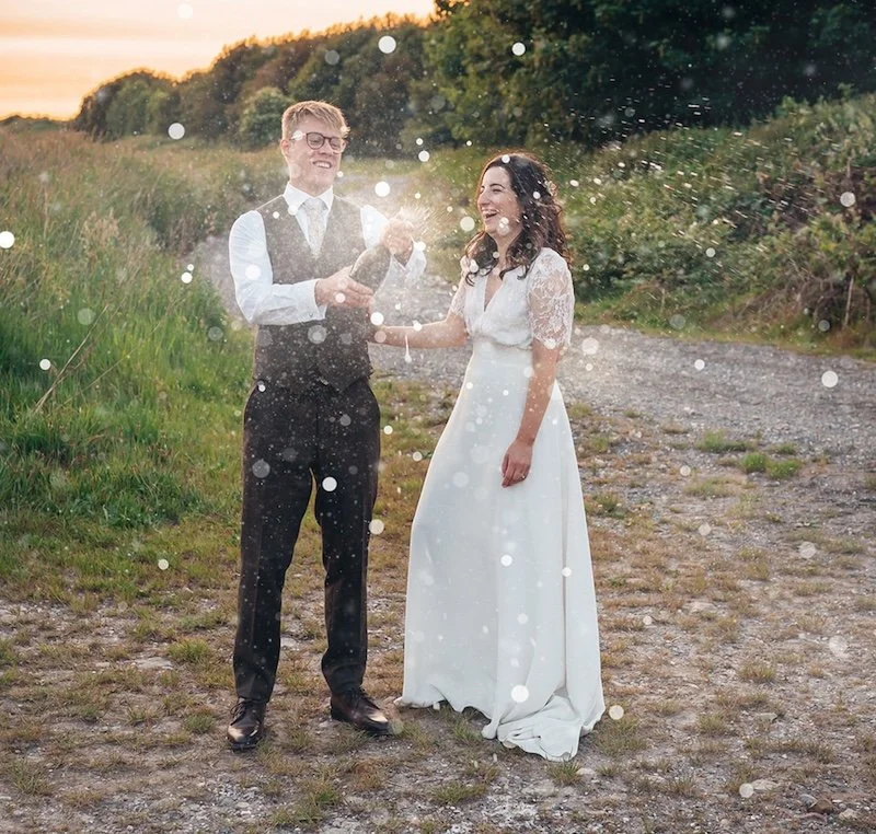 Couple celebrating their wedding by popping champagne in a beautiful field by their rustic barn wedding venue
