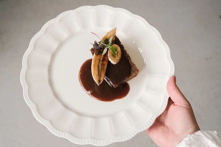 Hand holding a pretty white plate of delicious rum and banana cake by Wild Fork West