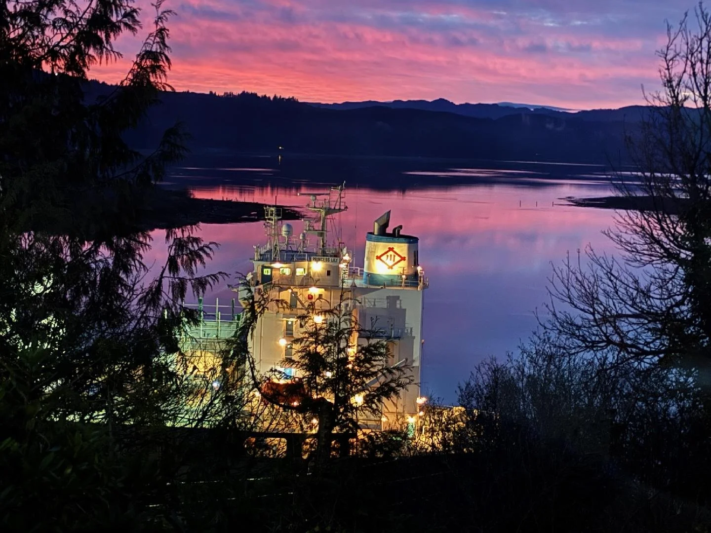 A ship near a calm body of water during sunset, with pink and purple clouds and mountain silhouettes in the background, surrounded by trees.