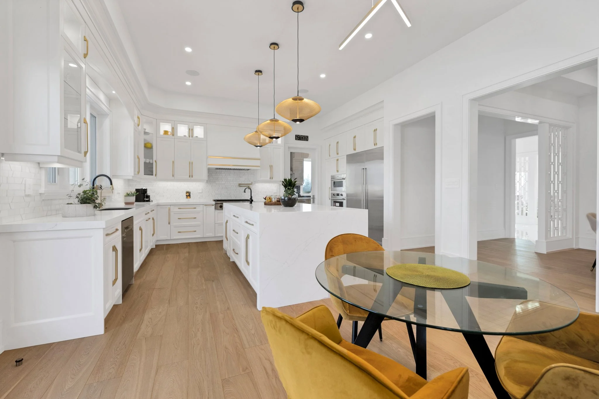 Modern white kitchen with gold accents, pendant lighting, and a glass dining table with yellow cushioned chairs.
