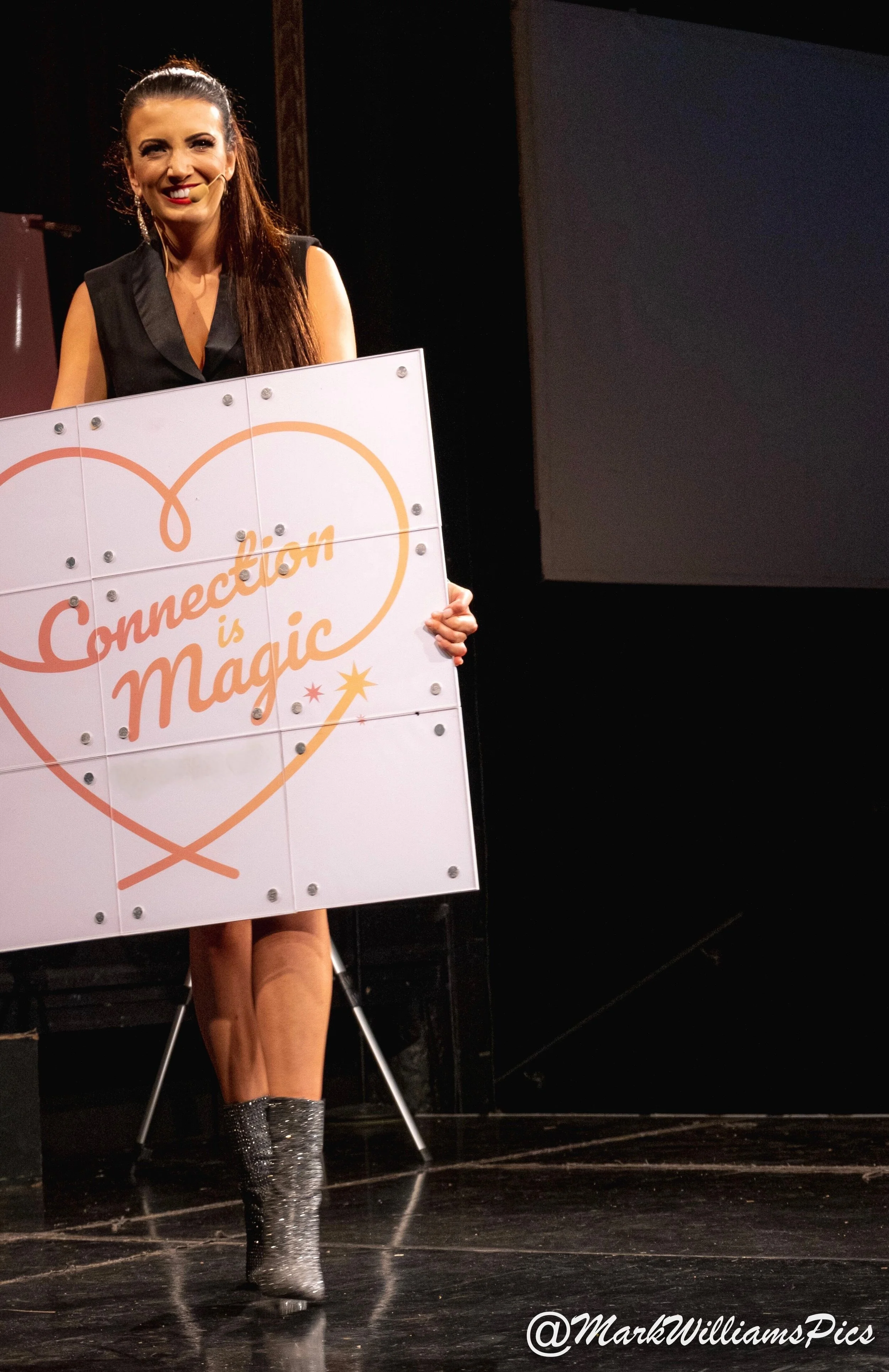 A woman on stage holding a sign that reads 'Connection is Magic' inside a heart outline. She is smiling, wearing a black sleeveless dress and sparkly knee-high boots, with a microphone headset. The background is dark, and the stage floor is black.