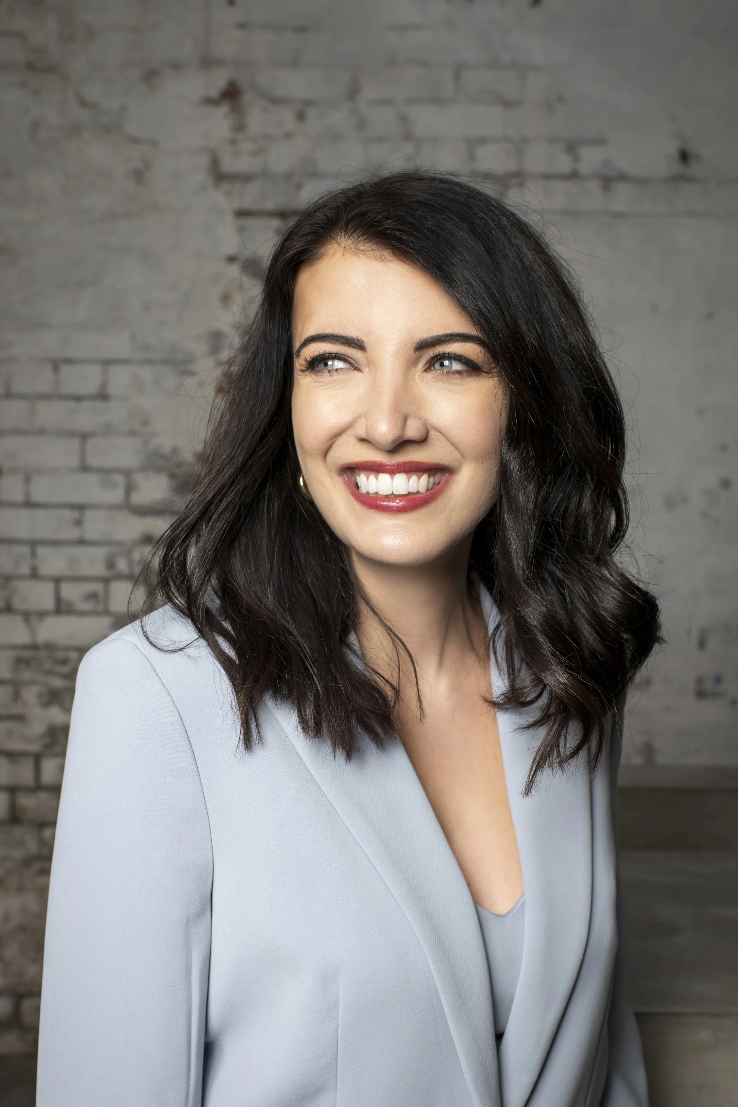 A smiling woman with dark, wavy hair wearing a light gray blazer and a matching top, standing against a textured gray brick wall background.