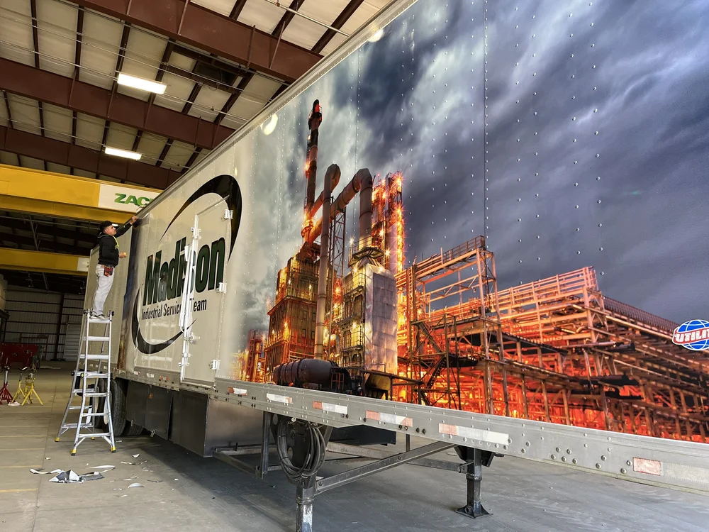 Person standing on a ladder applying a large industrial-themed decal to the side of a trailer inside a warehouse.