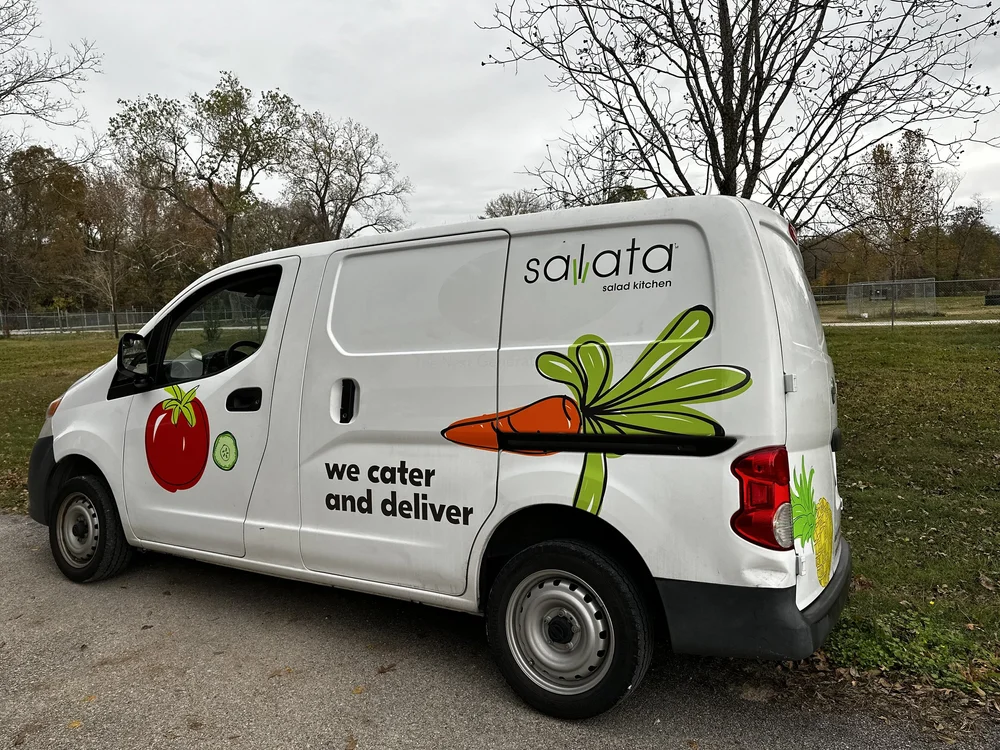 A white delivery van with Salata salad kitchen branding, featuring images of vegetables like tomatoes, cucumbers, carrots, and greens, and text saying "we cater and deliver."