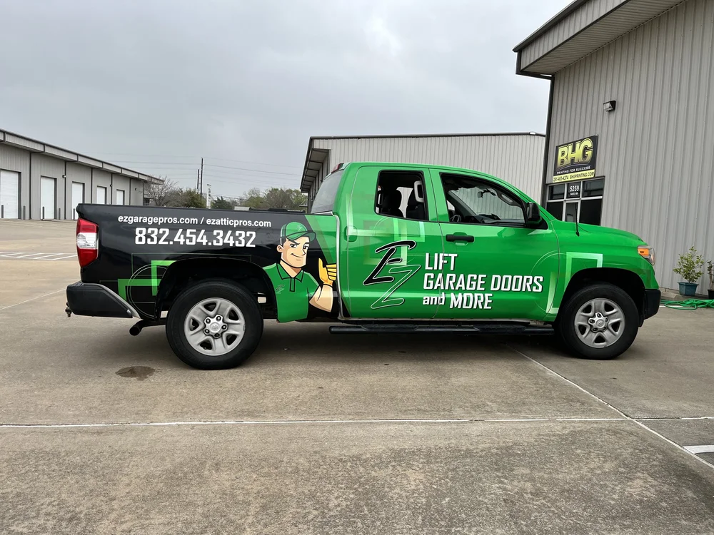 A green pickup truck with company branding for EZ Lift Garage Doors and More, featuring contact information and a cartoon character, parked in front of an industrial building.