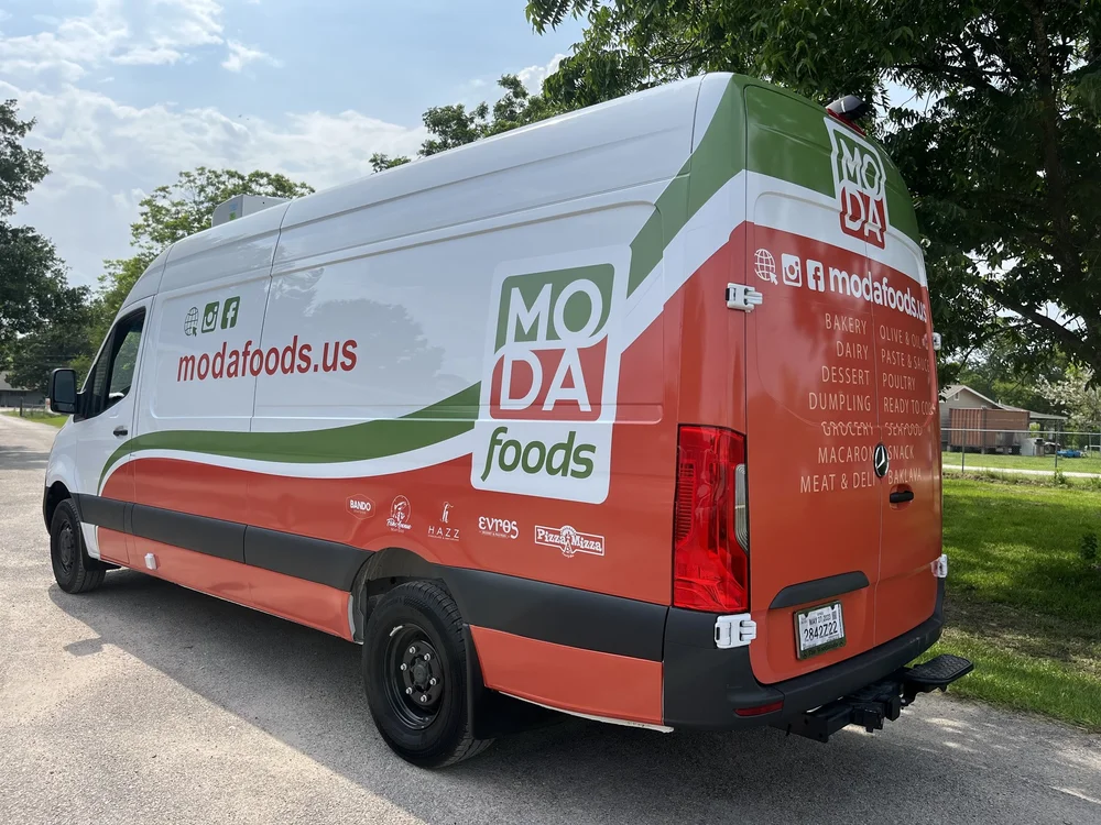 A delivery van for Moda Foods with branding and website details on the side, parked on a street. The van is white with green and orange accents, displaying food categories like bakery, dairy, dessert, and more. Trees and houses are visible in the bac