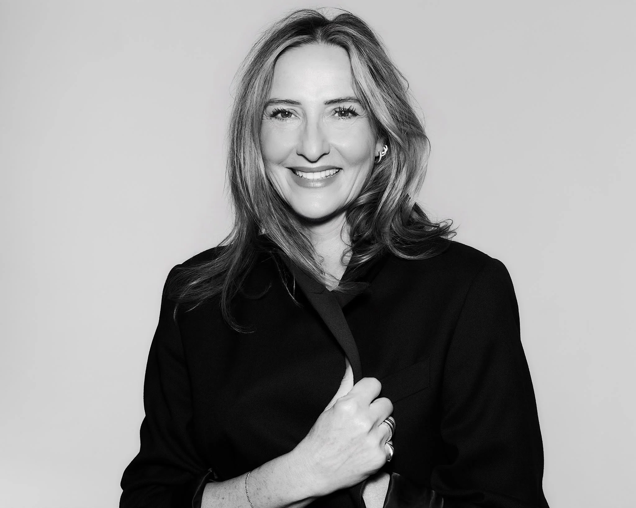 Black and white portrait of a smiling woman with wavy hair, wearing a dark top and a ring, standing against a plain background.