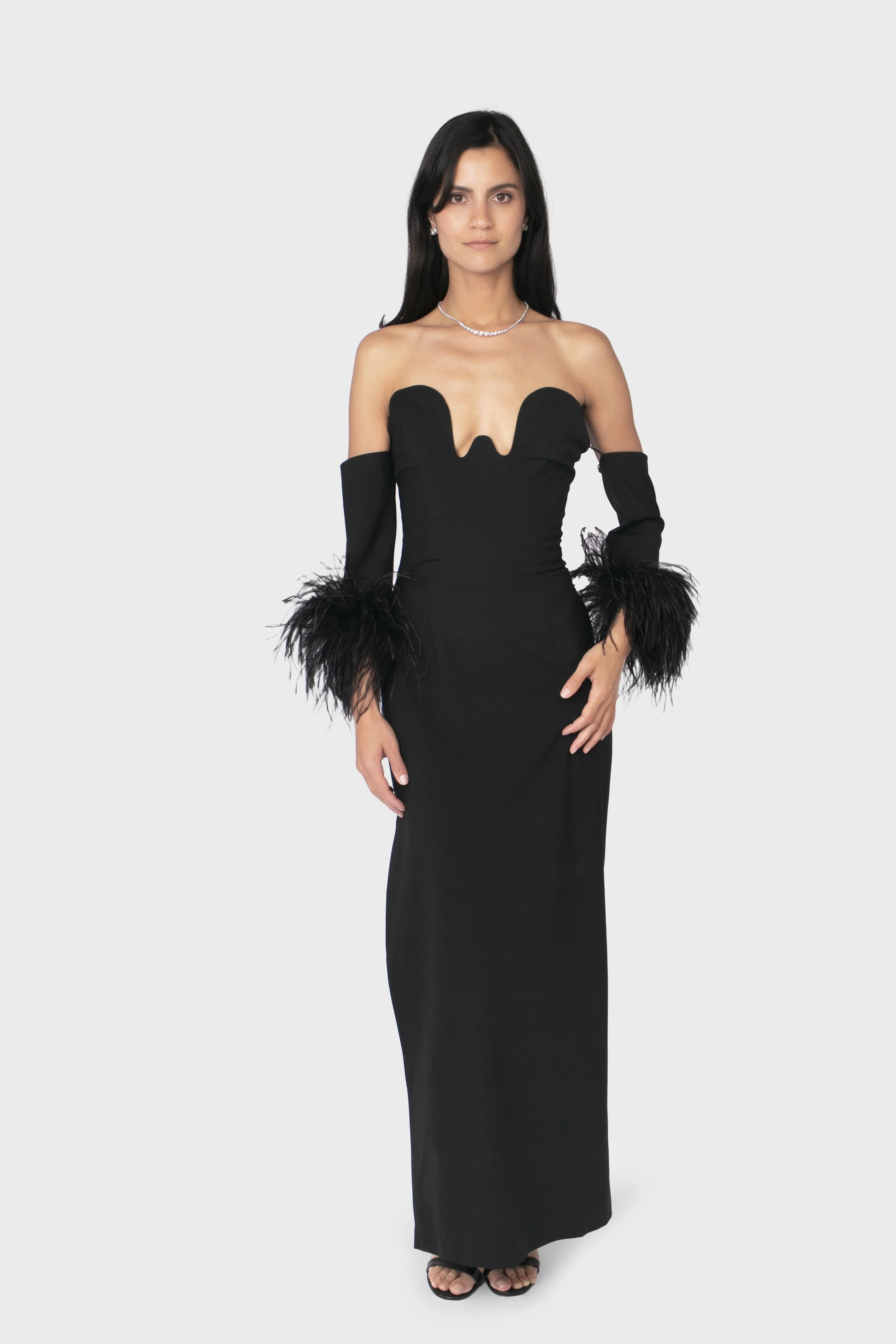Une femme porte une robe noire longue avec des détails en plumes sur les manches et un décolleté sophistiqué.
