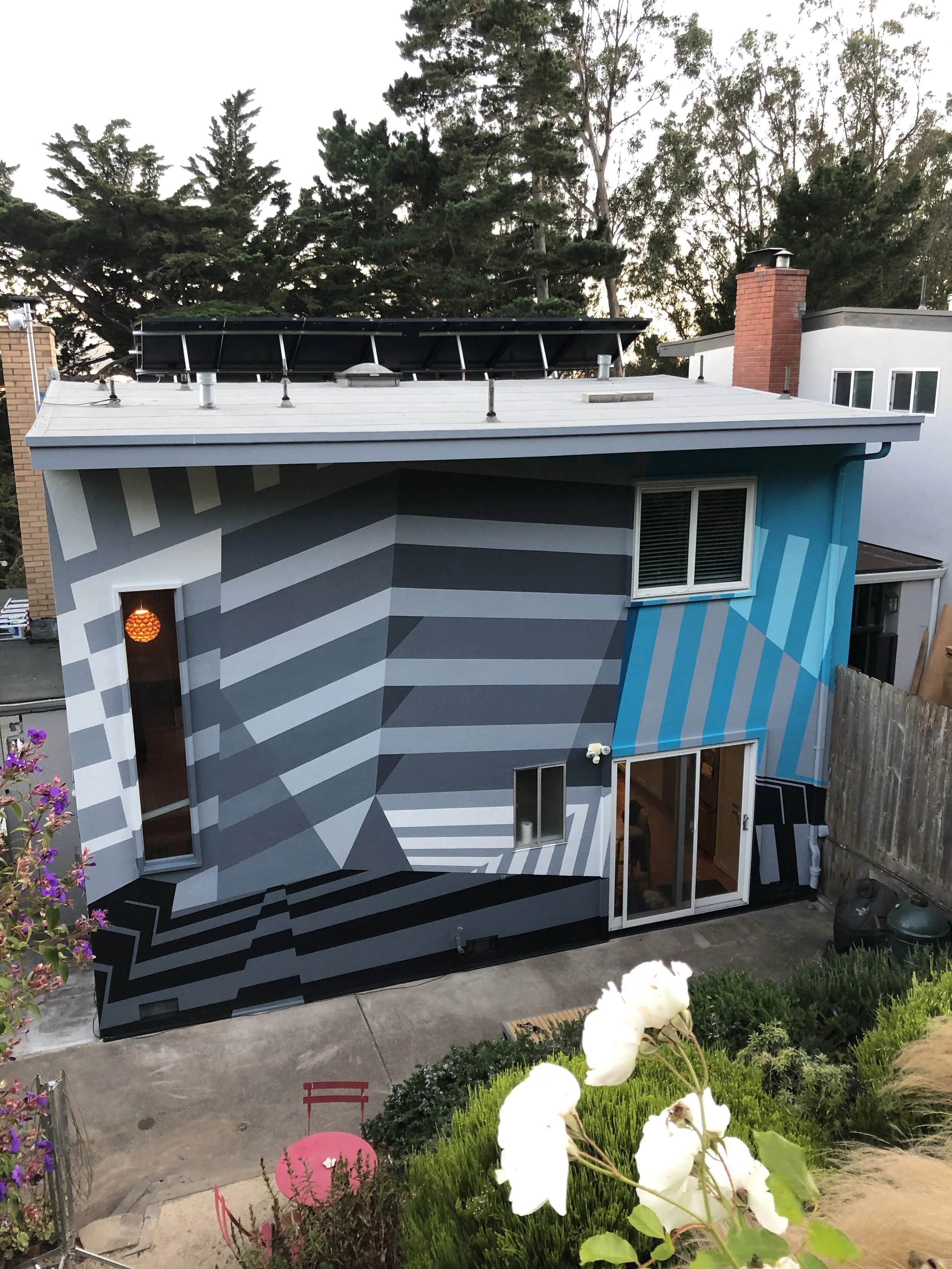 A modern house with geometric black, white, and blue mural on the exterior wall, featuring a sliding glass door, window, and a small outdoor table with chairs, surrounded by greenery and flowers.