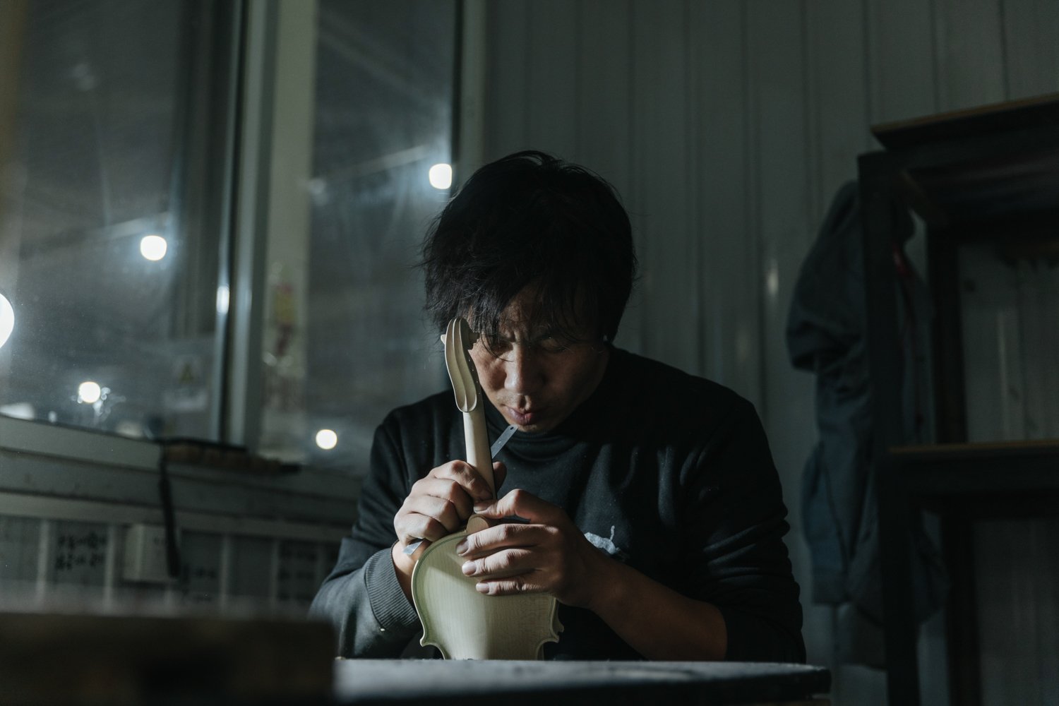 Ein junger Mann, der an einem Tisch in einem Raum sitzt, konzentriert ein Musikinstrument, eine Geige, zu reparieren oder zu bauen. Es ist dunkel, und das Licht kommt von einer großen Scheibe im Hintergrund.