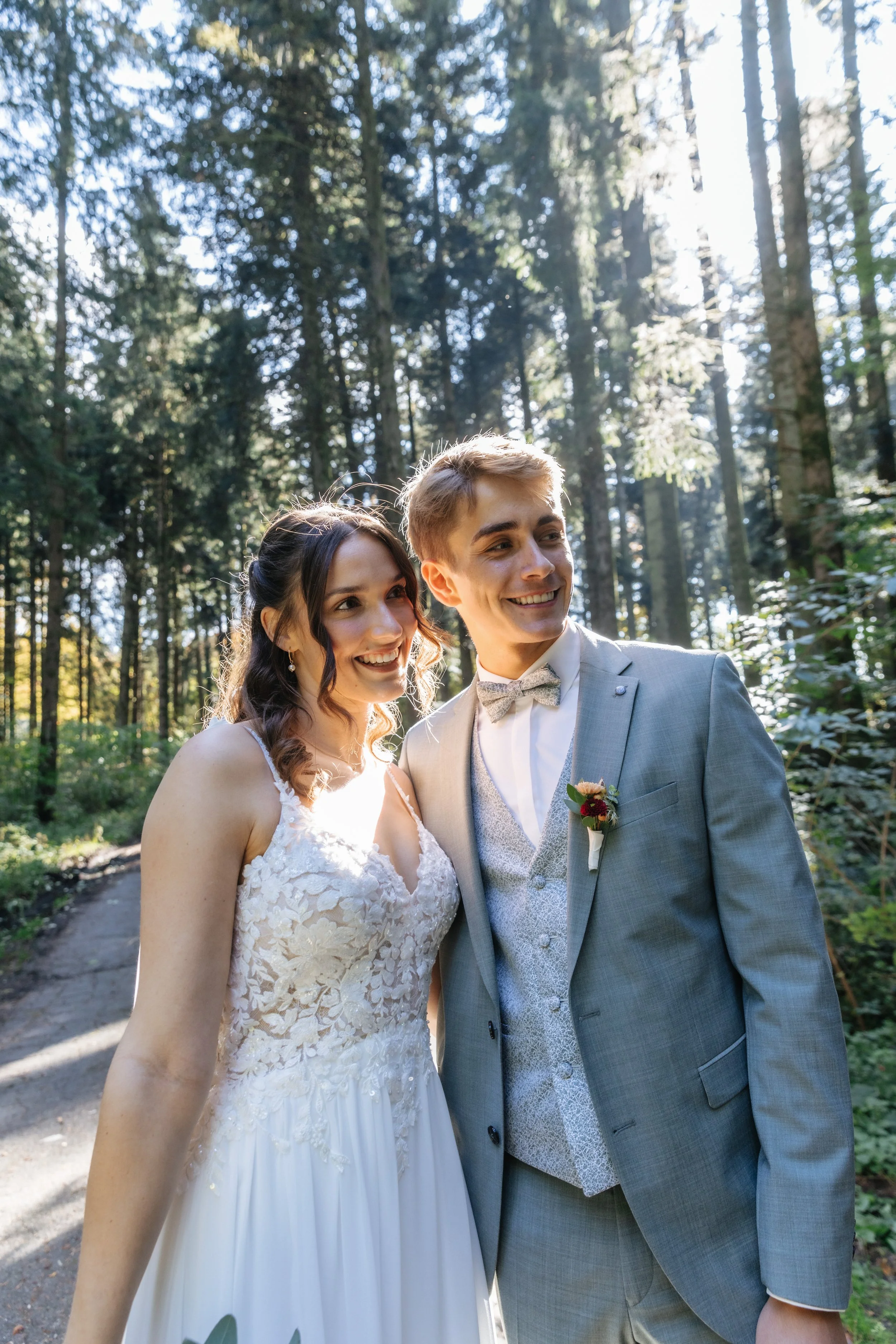 Ein glückliches Brautpaar in Hochzeitkleidung steht im Wald und lächelt.