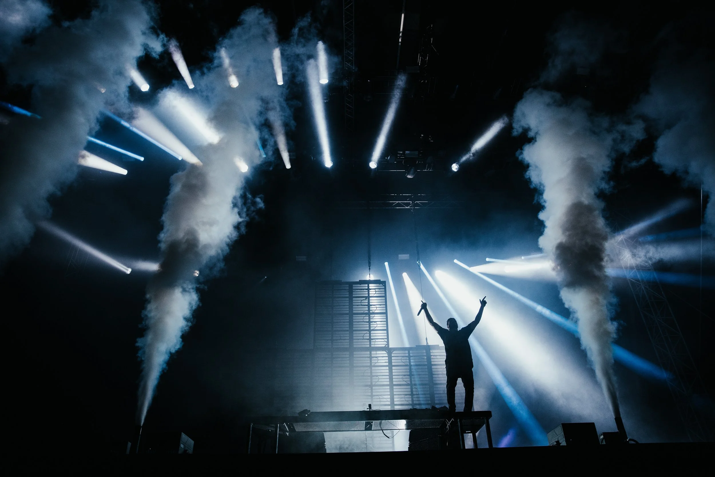 Ein Sänger auf einer Bühne, umgeben von weißen Rauch- und Lichteffekten, während er mit ausgestreckten Armen auf einem Podest steht.