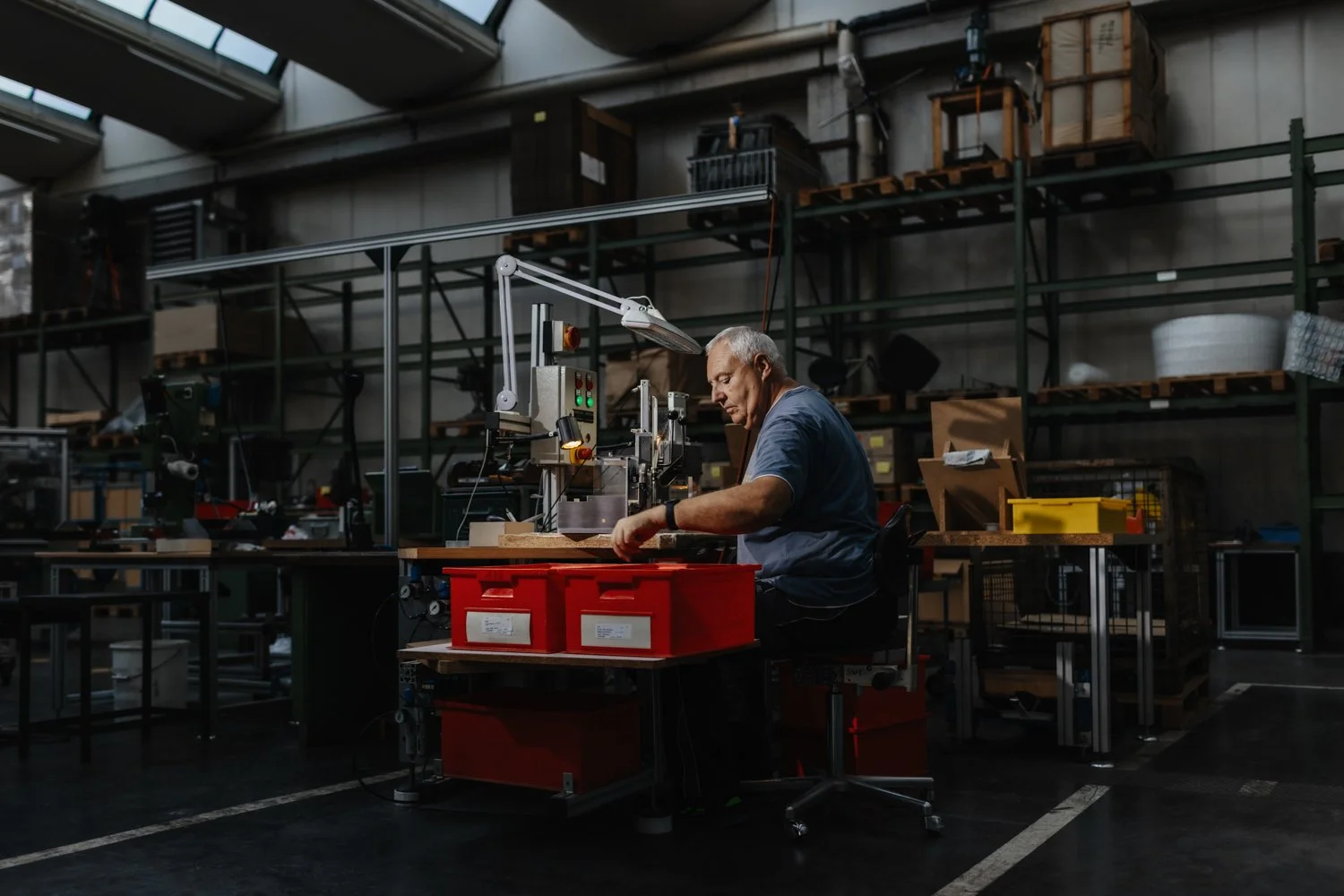 Ein älterer Mann arbeitet in einer Werkstatt mit Maschinen und Werkzeugen, umgeben von verschiedenen Lagerregalen und Kartons.