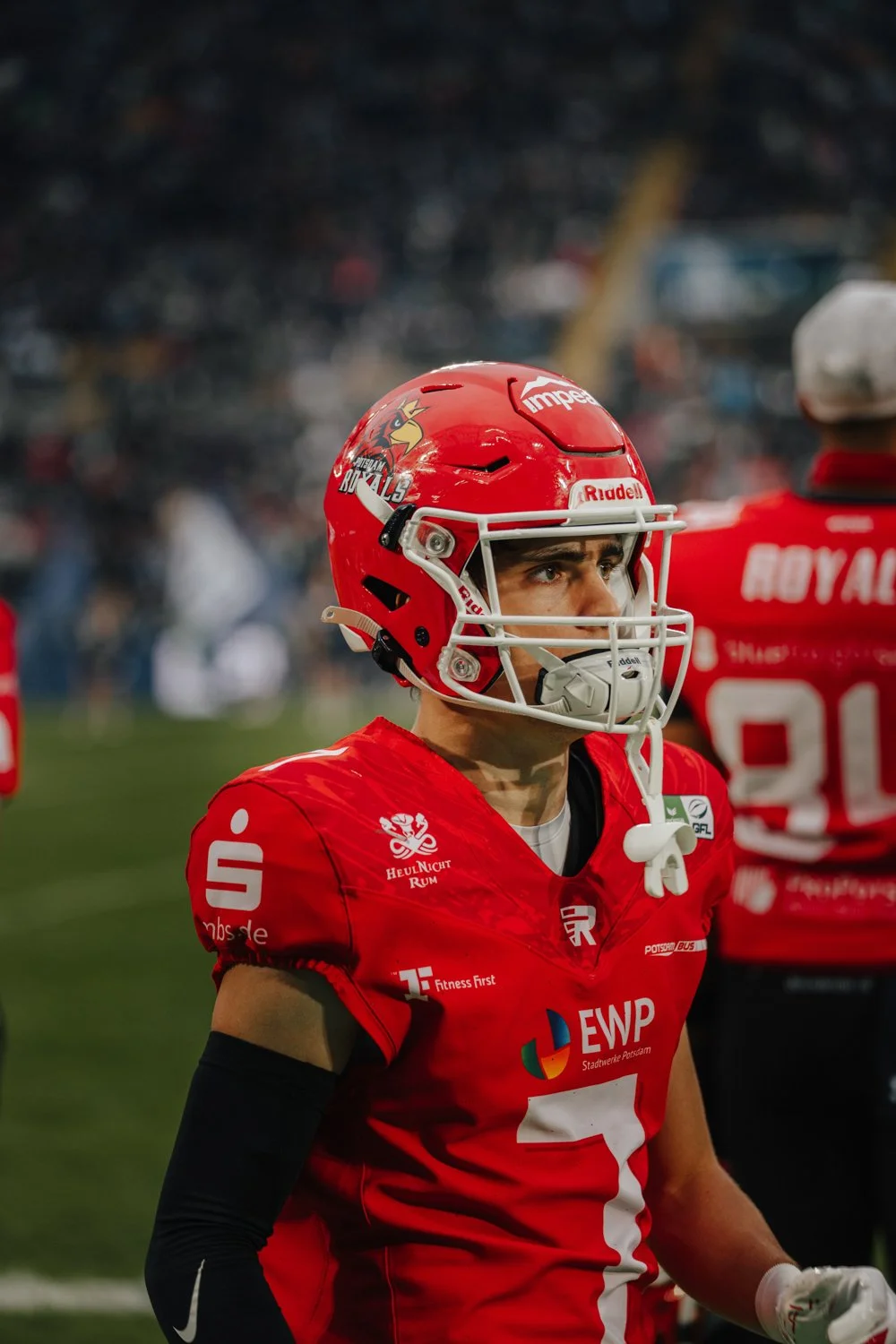 Footballspieler in roter Uniform und Helm auf dem Spielfeld mit Blick nach rechts, im Hintergrund andere Spieler und Zuschauer im Stadion.