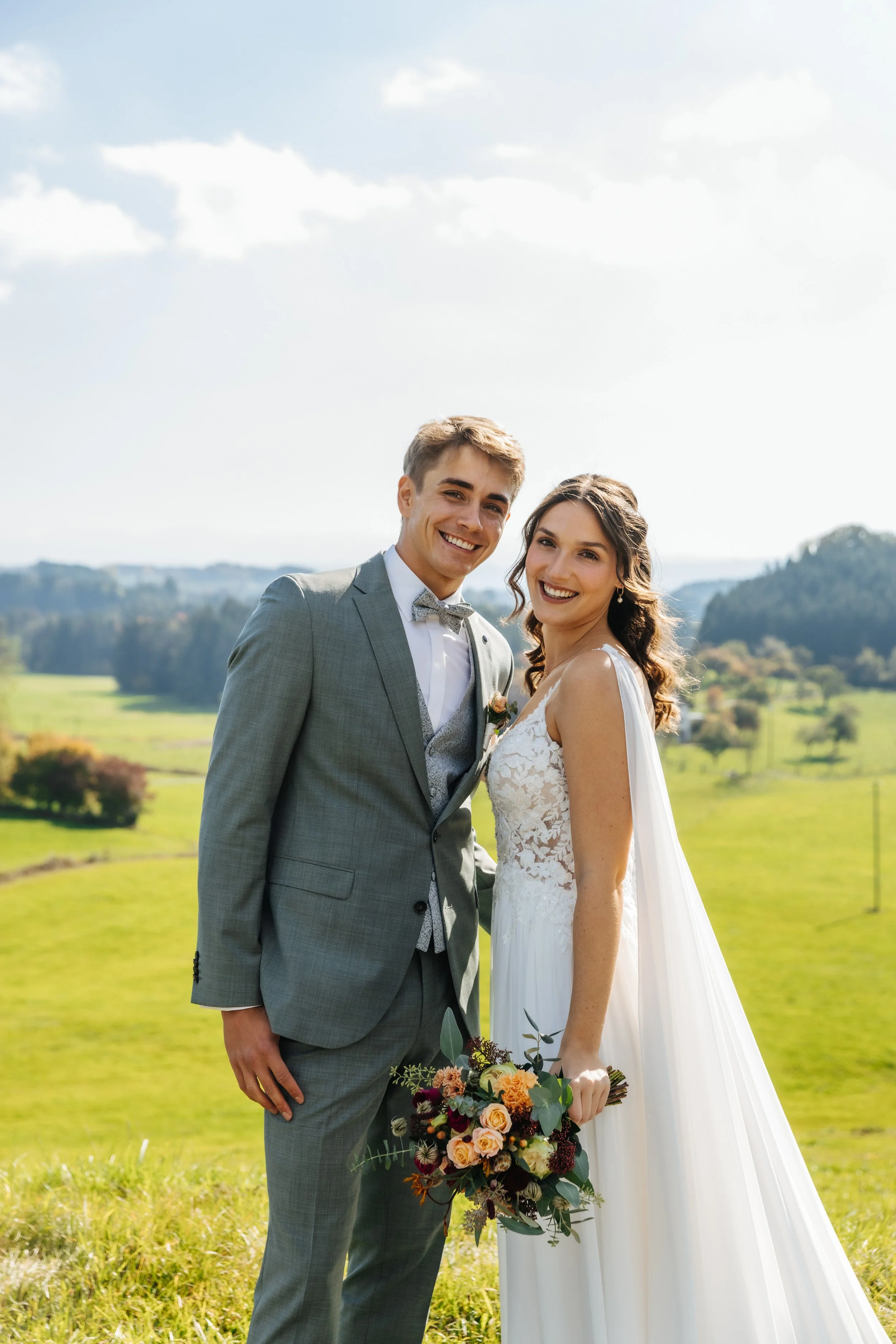 Ein frisch verheiratetes Paar steht in einer grünen Landschaft bei sonnigem Wetter und lächelt. Der Bräutigam trägt einen grauen Anzug mit Fliege, die Braut ein weißes Hochzeitskleid und hält einen Blumenstrauß.