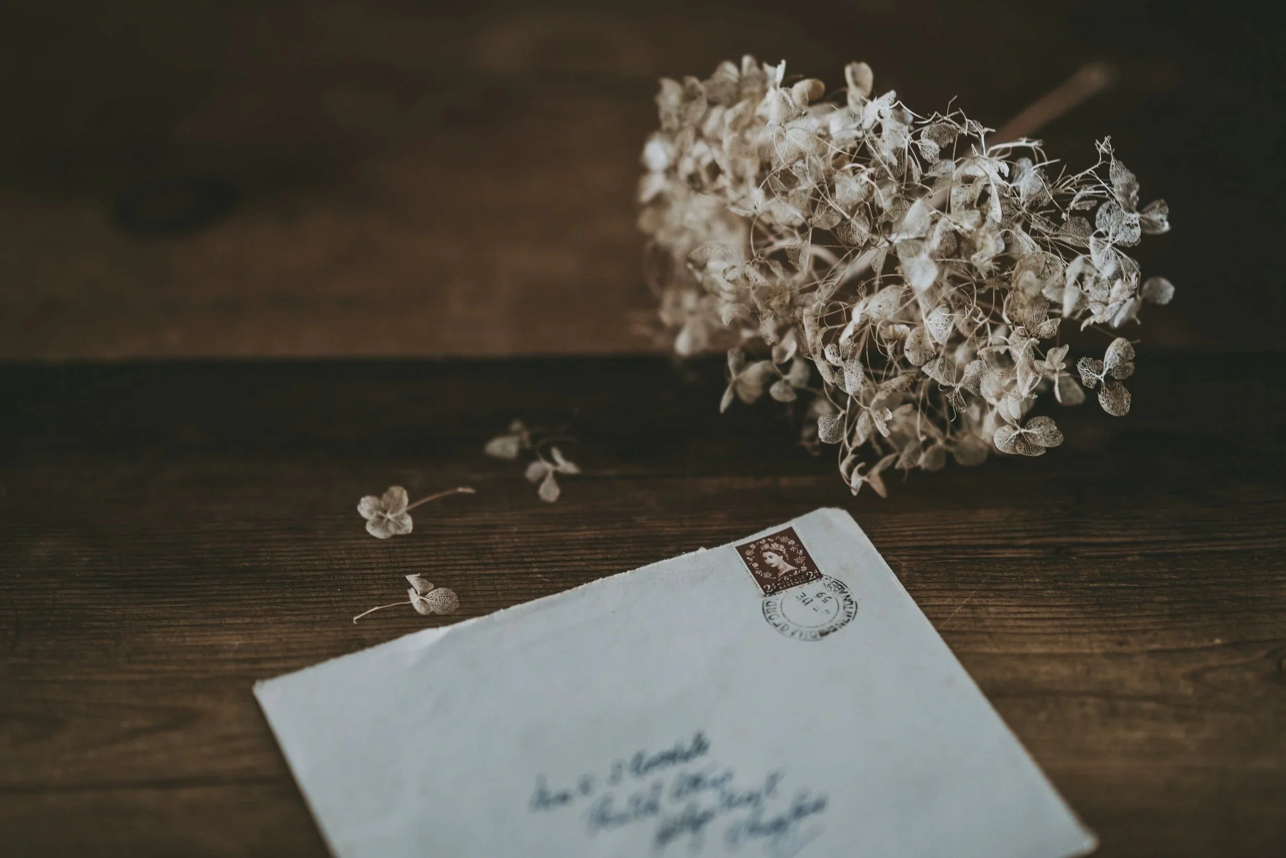 A dried bouquet of flowers resting on a wooden surface next to a handwritten envelope with a postage stamp.
