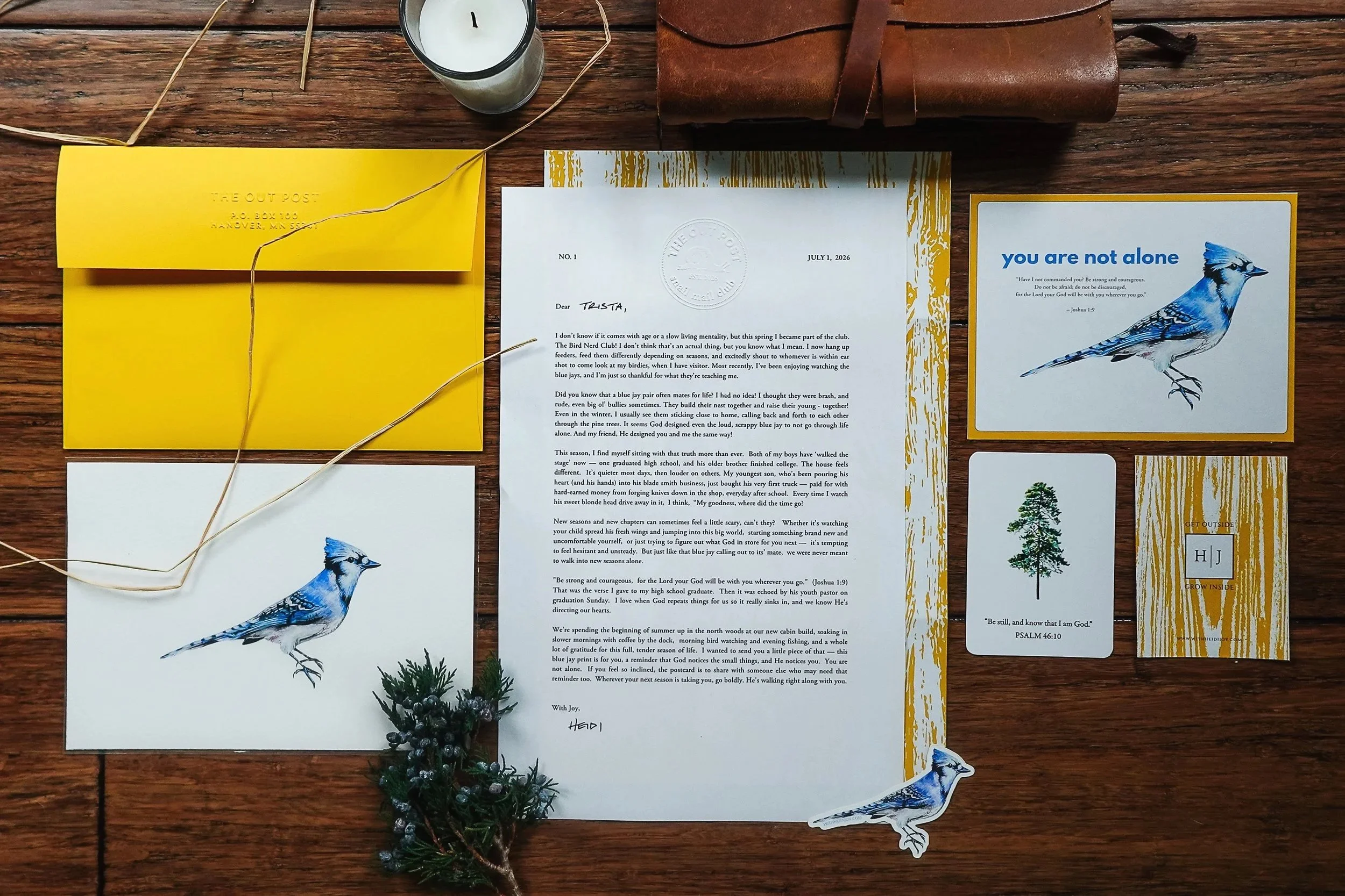 A flat lay of a wooden table with various items. There are yellow and white cards, a letter with a date of July 1, 2026, a small decorative pine arrangement, a bird sticker, a black and white candle, and a rolled piece of leather.