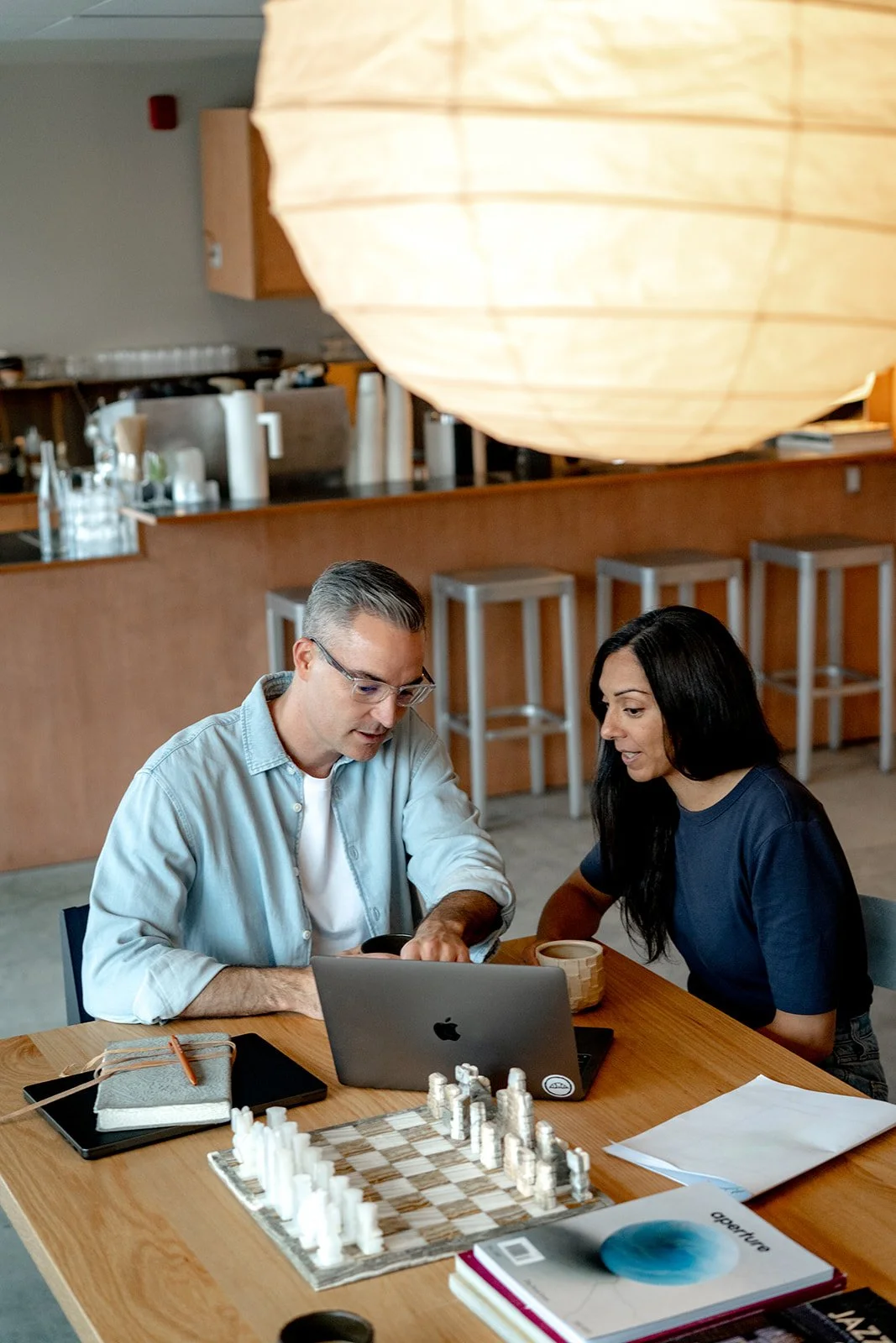 Modern workspace with a silver laptop, black smartphone, wooden desk lamp, white cup, and a black napkin on a white desk, with a large window in the background.