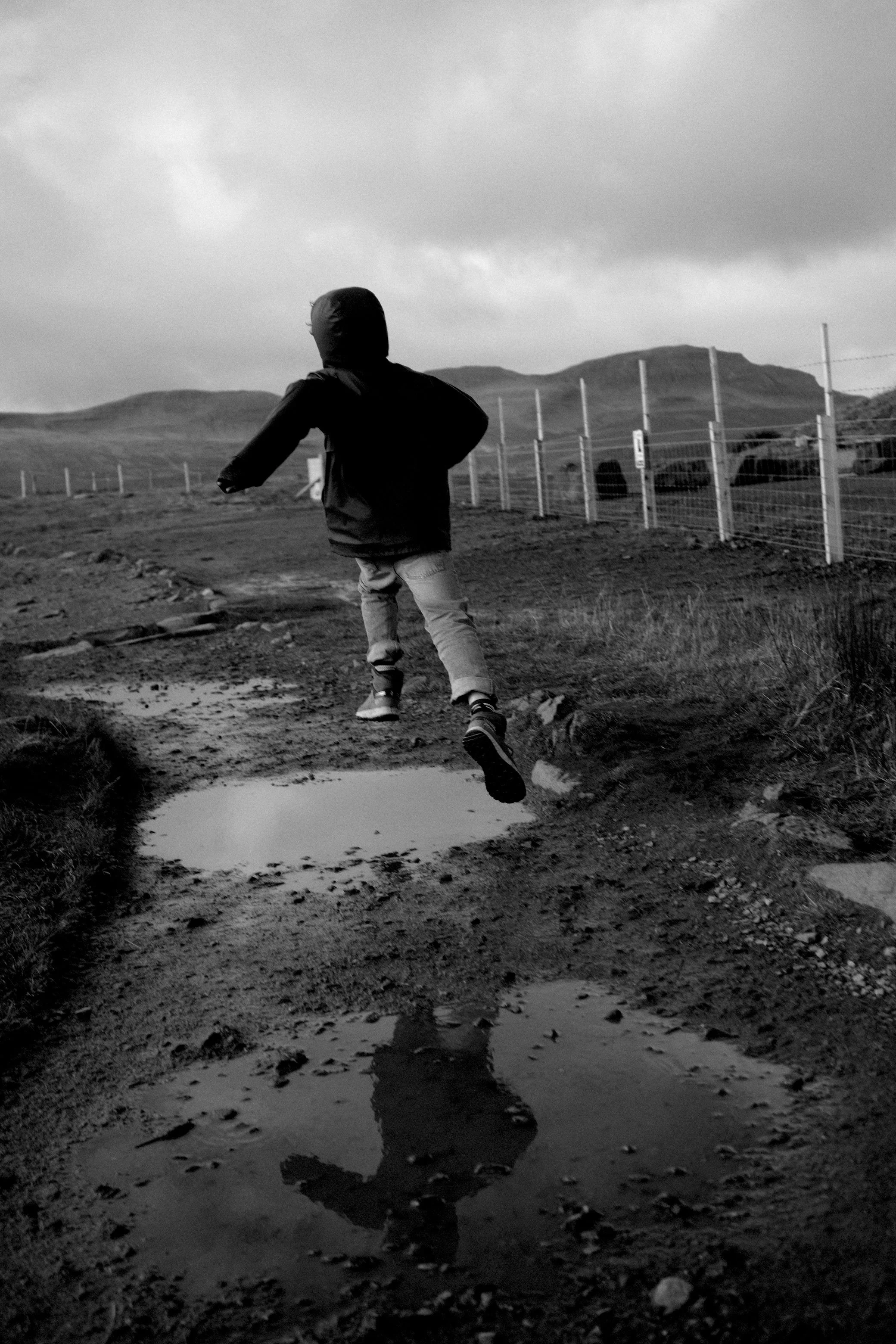 Un enfant court sur un chemin boueux avec des flaques d'eau, sous un ciel nuageux, avec des montagnes en arrière-plan.