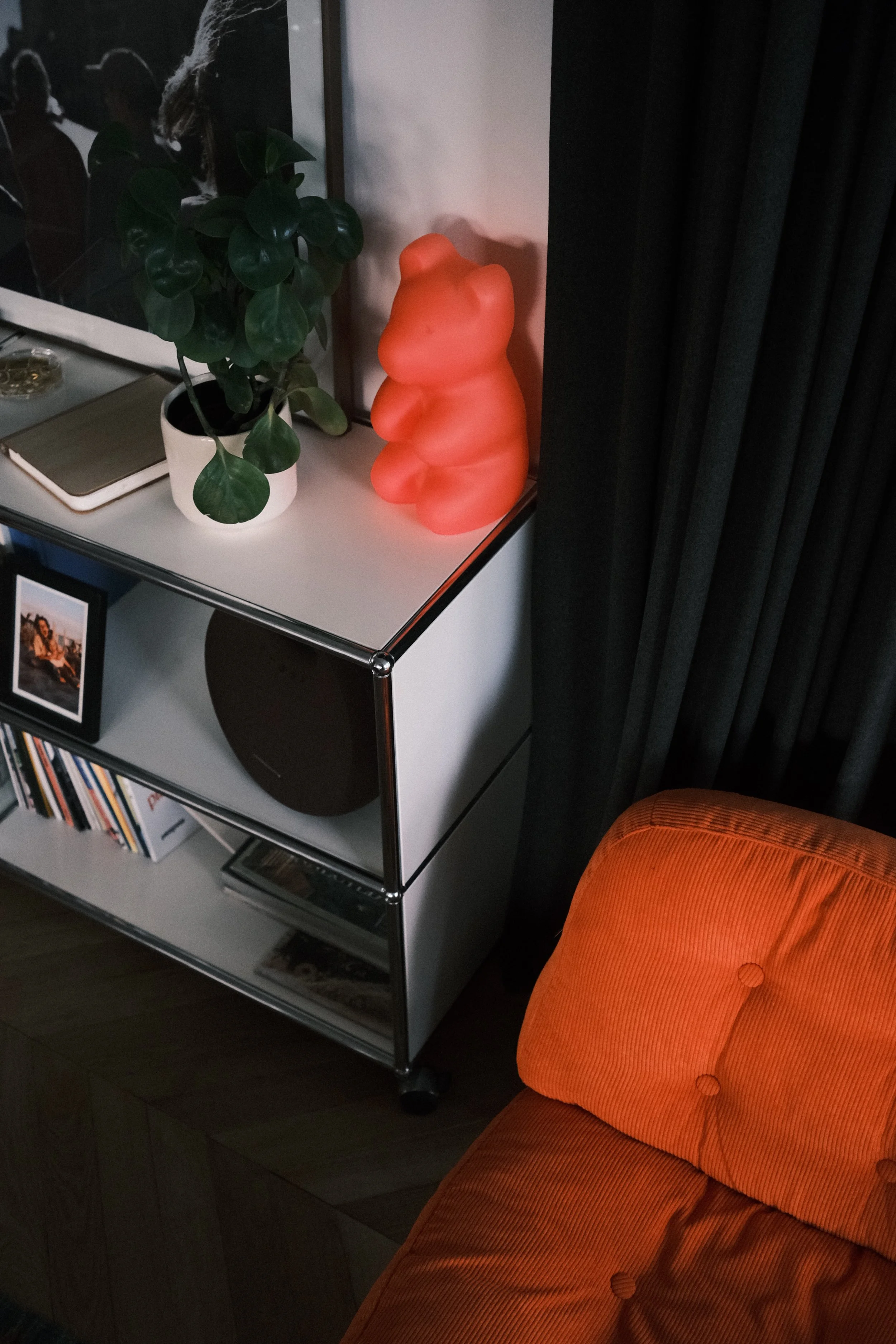Une étagère en métal avec un pot de plante verte, une lampe en forme de ourson rose et une photo encadrée en dessous, à côté d'un fauteuil orange à coussin.