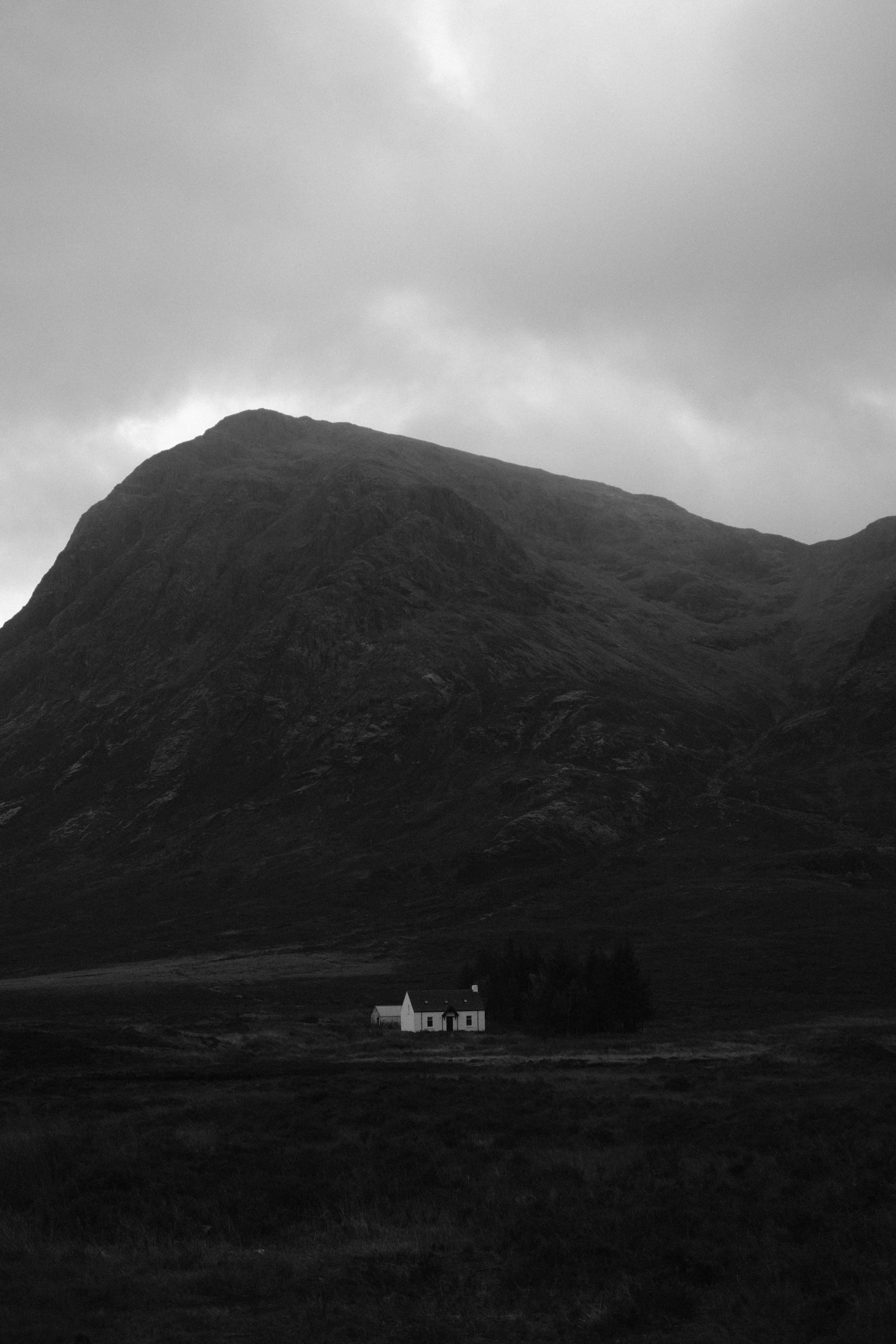 Photographie en noir et blanc d'une petite maison blanche entourée d'arbres, située dans une vallée avec une grande montagne en arrière-plan sous un ciel nuageux.