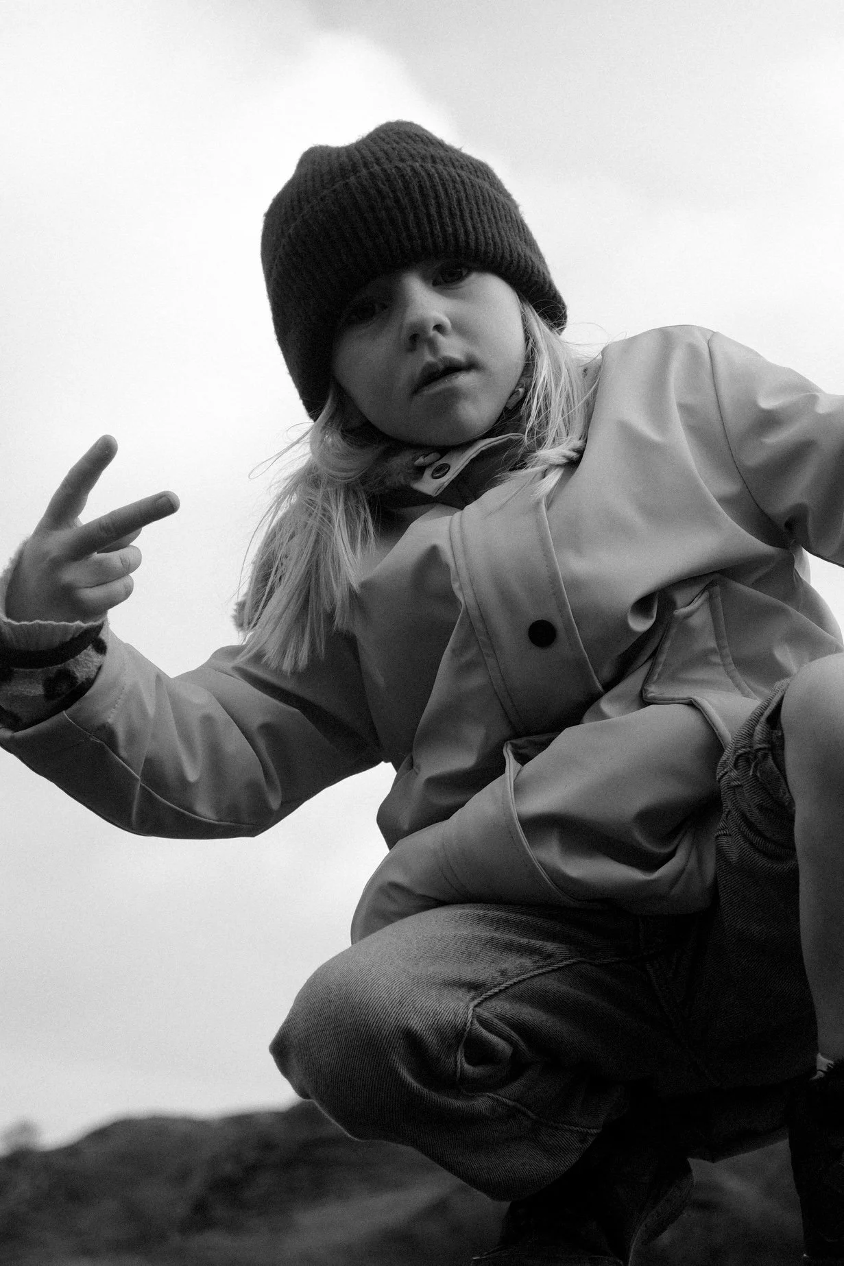 Une jeune fille avec un bonnet noir, portant une veste, fait un signe de paix avec deux doigts. Elle se trouve en extérieur, dans un paysage qui semble rocheux ou montagneux.