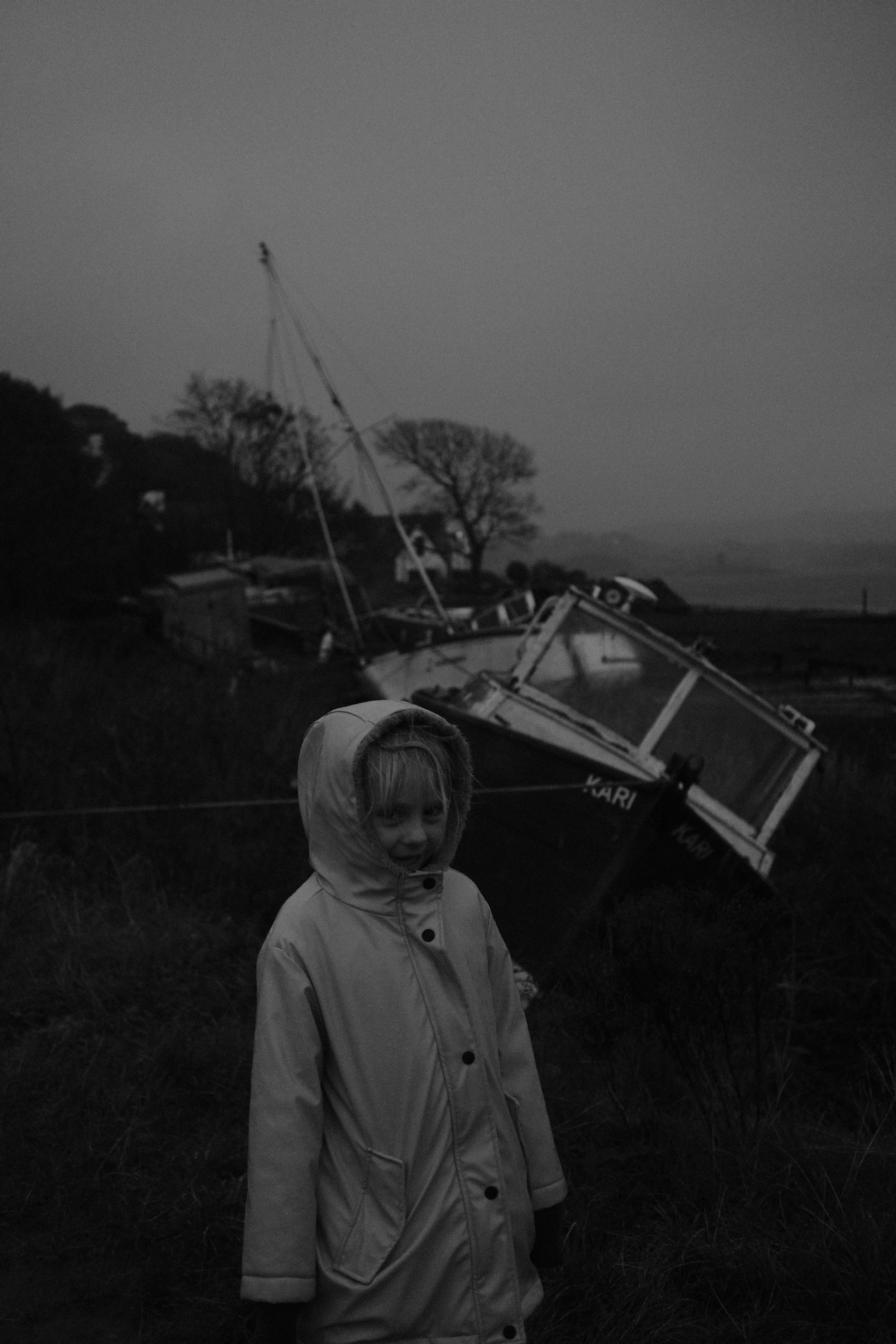Un garçon en manteau avec capuche devant un bateau renversé sur la rive, le ciel est gris.