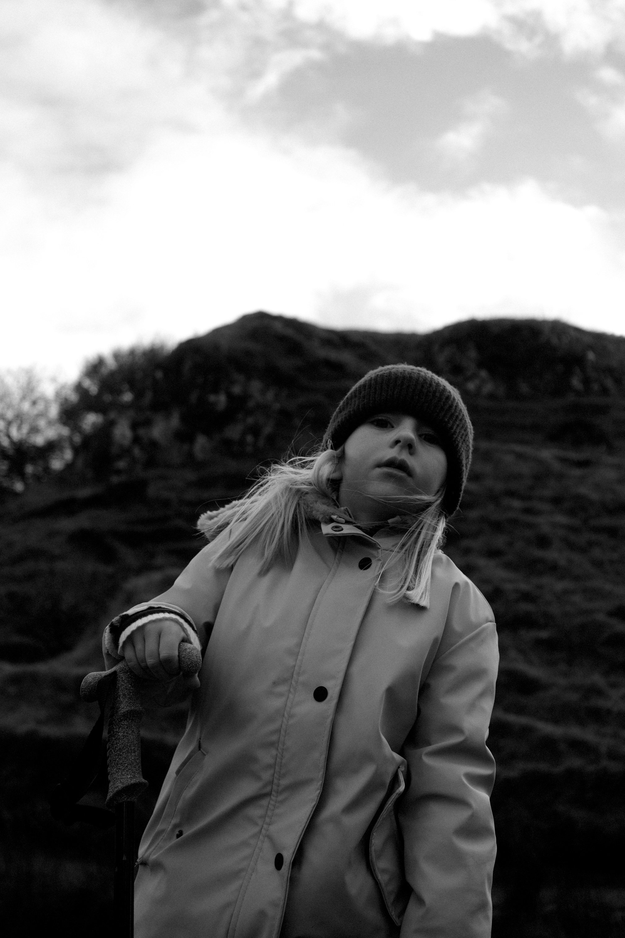 Une fille portant un manteau et un bonnet, tenant une canne, en extérieur avec des collines verdoyantes en arrière-plan, en noir et blanc.